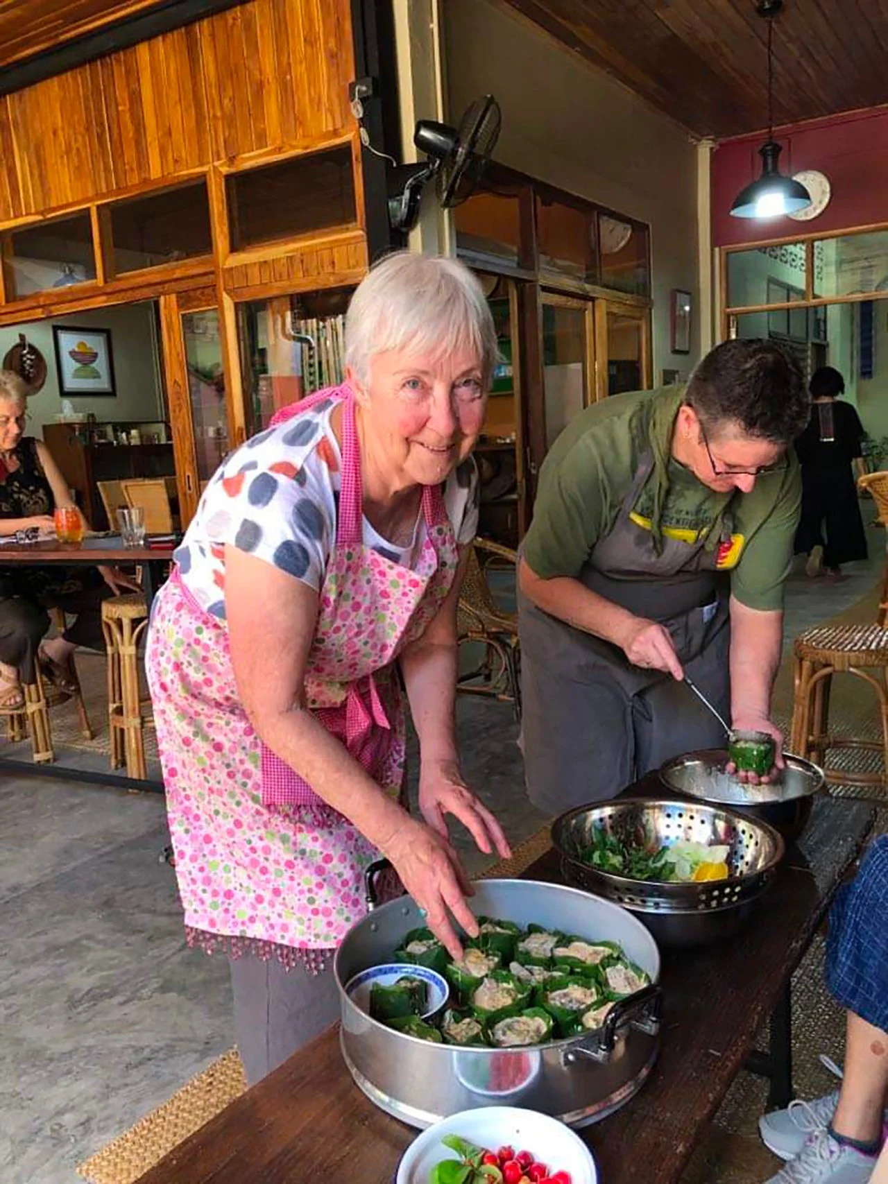 A woman and man preparing food in a restaurant, with the woman placing tray with stuffed green peppers, and the man peeling or slicing green peppers.