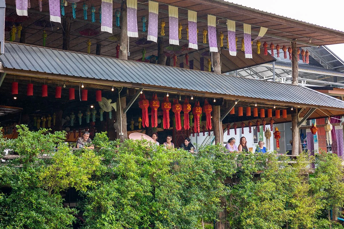 People sitting on a balcony decorated with colorful lanterns and hanging ornaments, surrounded by lush green bushes.