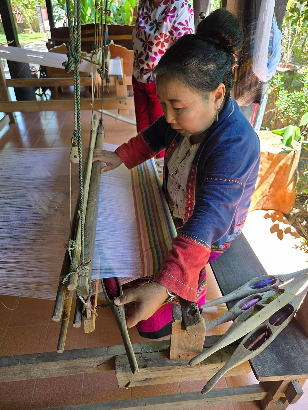 A woman dressed in traditional attire weaving on a loom, surrounded by a bright, outdoor setting with plants and sunlight streaming in.