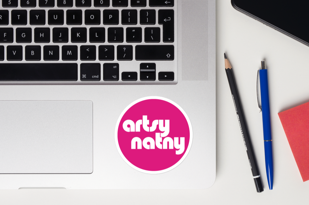 Close-up of a silver laptop keyboard with a pink circular sticker that says "artsy natny" on the laptop's surface, next to a black pencil, blue pen, red notepad, and part of a black smartphone on a white desk.