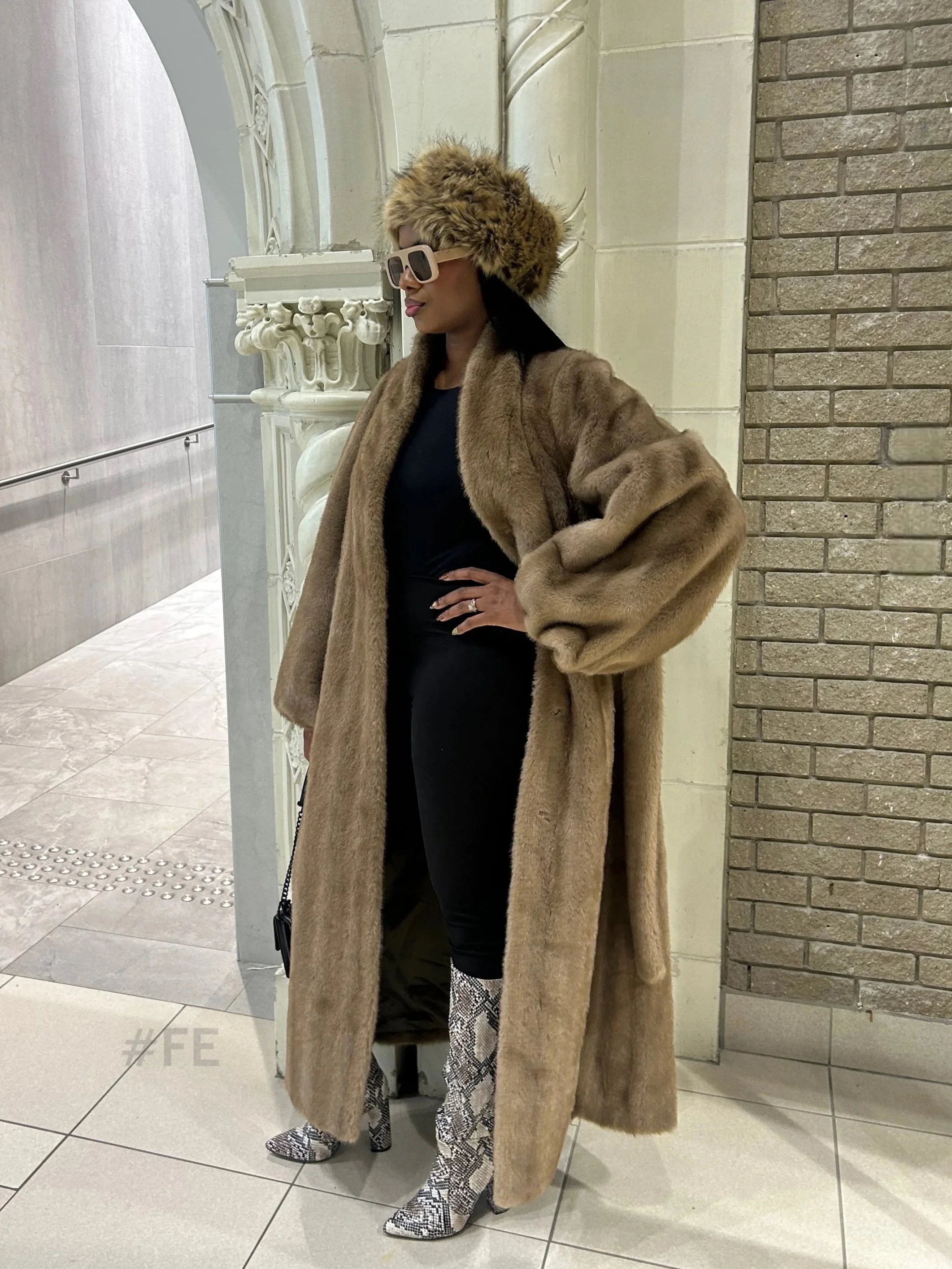 Une femme portant un manteau en fourrure beige, un chapeau en fourrure, des lunettes de soleil, des bottes à motifs python, et posant dans un intérieur élégant avec des murs en pierre et en plâtre.