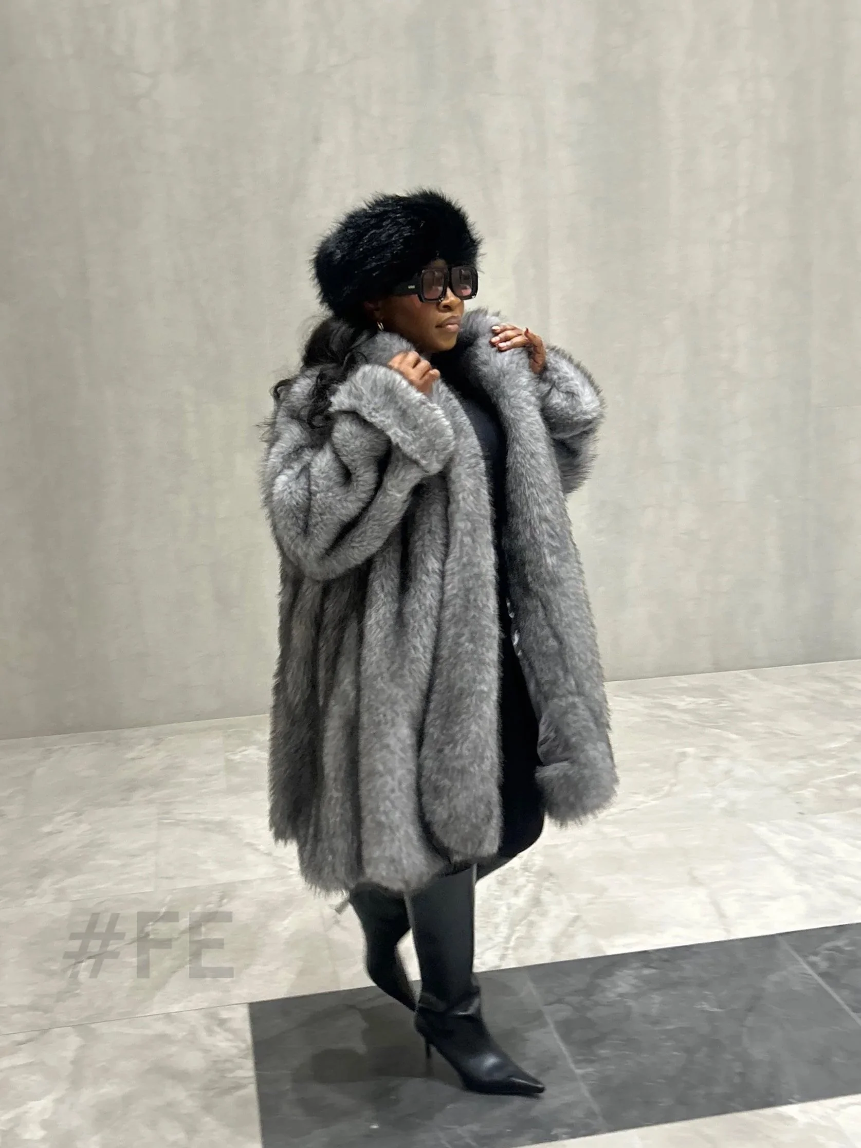 Une femme élégante portant un manteau en fourrure gris, un chapeau en fourrure noir, des bottes en cuir noir et des lunettes noires, posant dans un décor intérieur moderne.