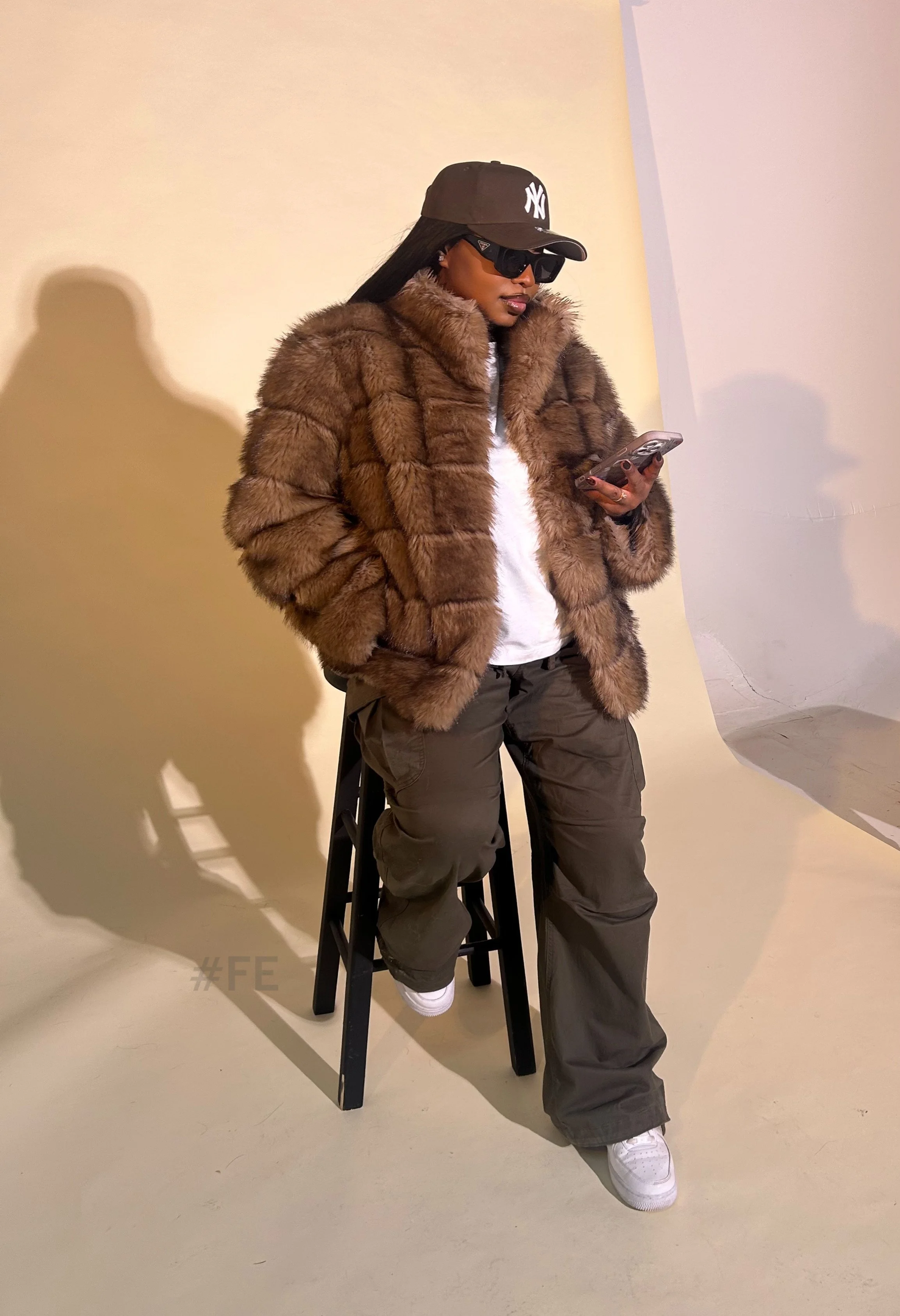 Une femme portant un manteau en fourrure brune, un chapeau gris avec le logo des Yankees, des lunettes de soleil noires, un t-shirt blanc, un pantalon cargo gris et des chaussures blanches. Elle est assise sur un tabouret noir en regardant son téléphone, dans un décor intérieur avec un mur beige clair et une lumière douce.