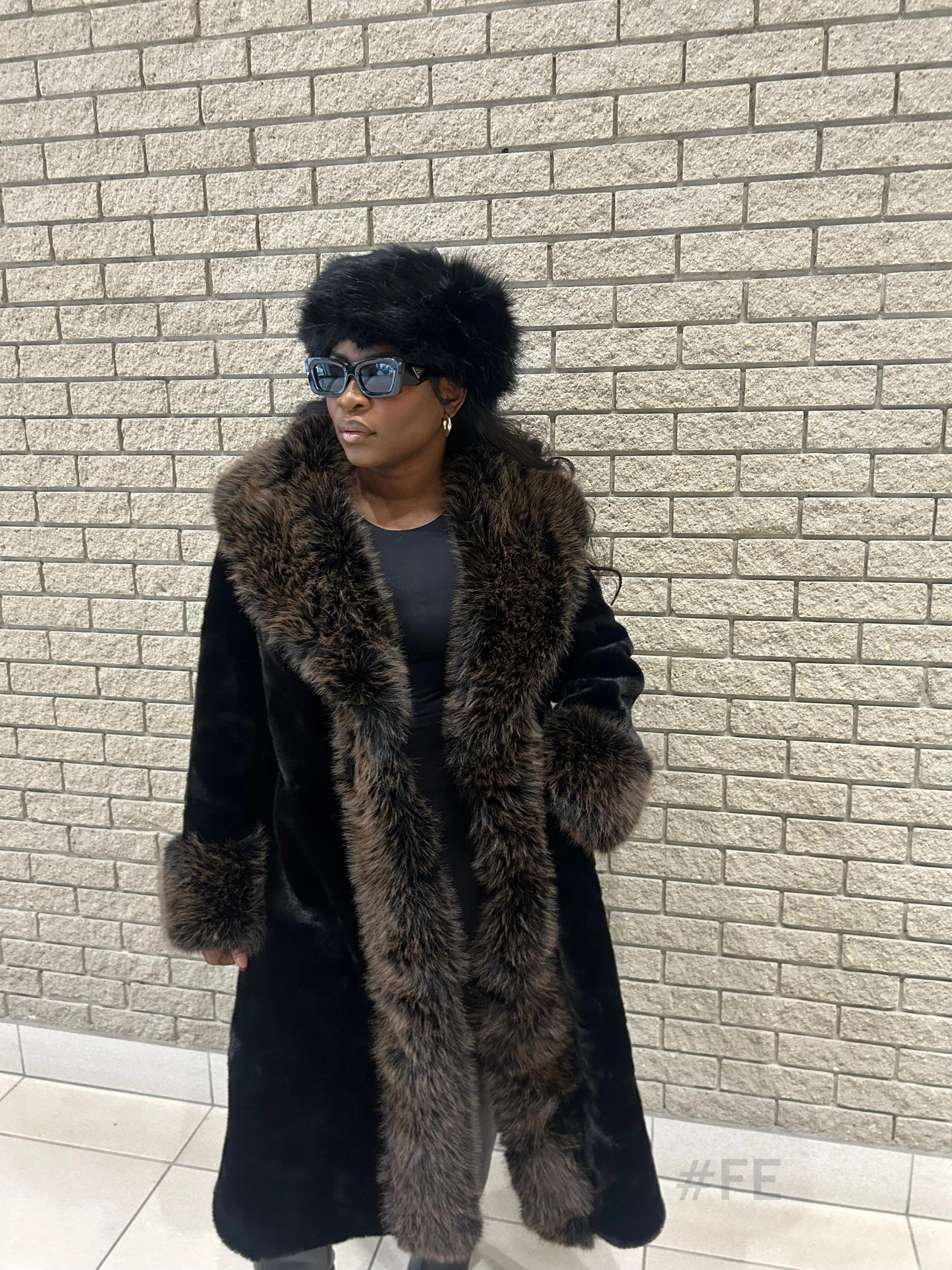 Femme portant un manteau en fourrure noire avec une longue bordure en fourrure marron, des lunettes de soleil noires, et un chapeau en fourrure noire, debout contre un mur en briques beige.