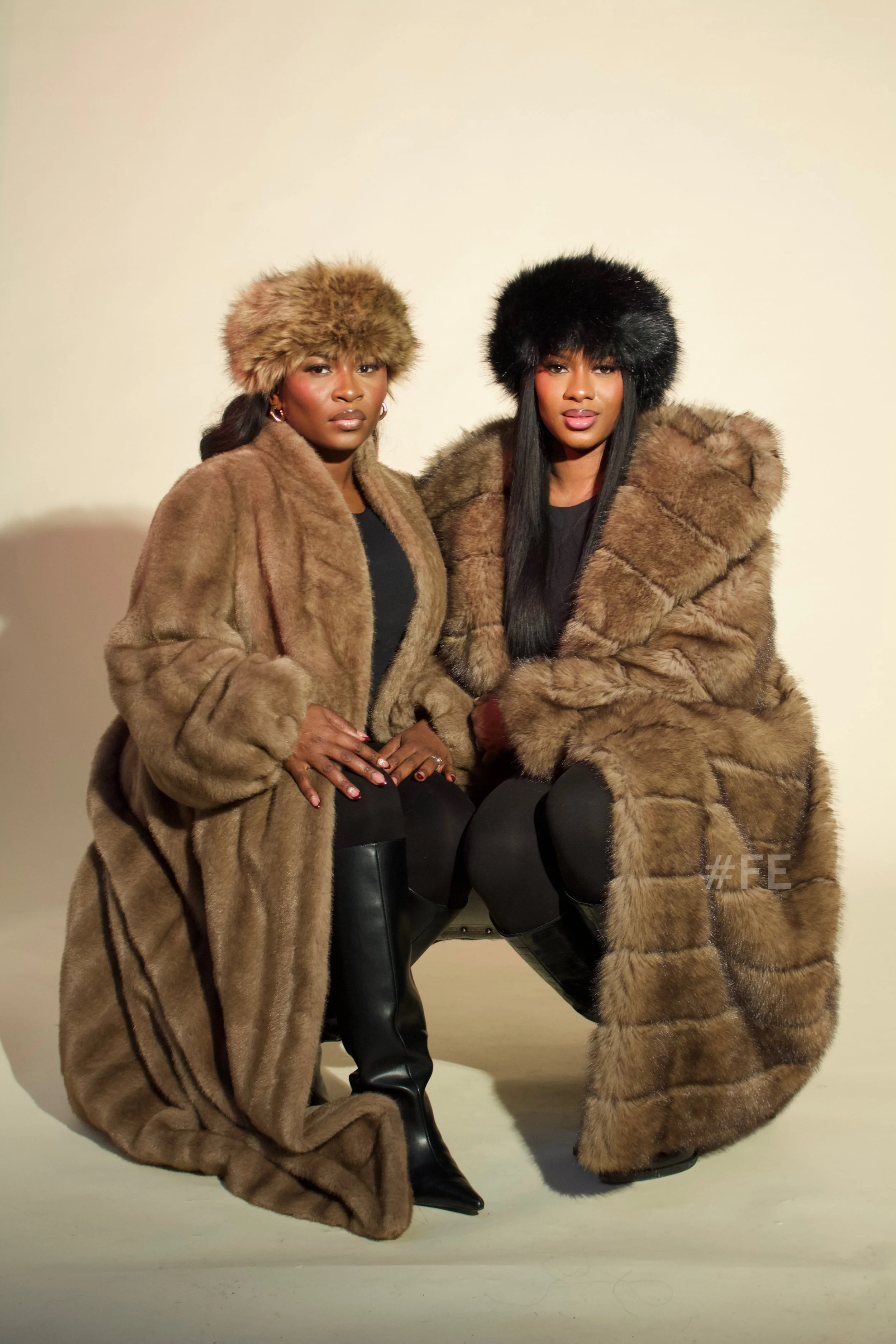 Deux femmes portant de longs manteaux en fourrure et des bonnets en fourrure, assises, contre un fond neutre.