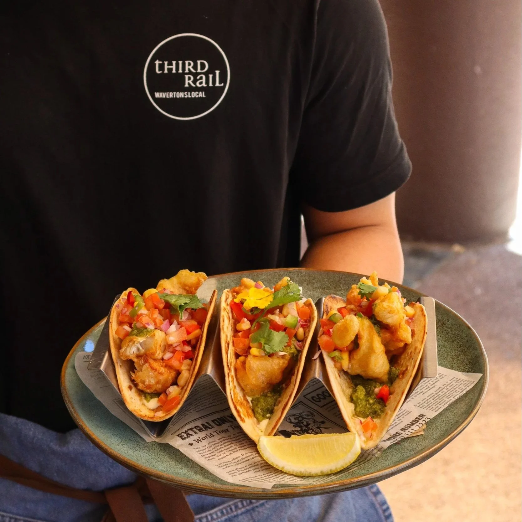 A person holding a plate with three tacos filled with fried fish, diced vegetables, cilantro, and topped with a slice of lemon.