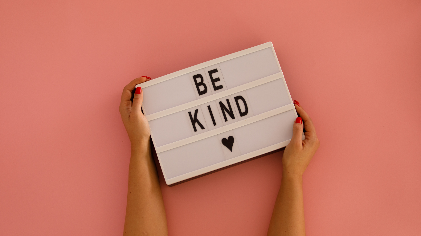 Person holding a white lightbox with the message 'BE KIND' and a heart symbol, against a pink background.