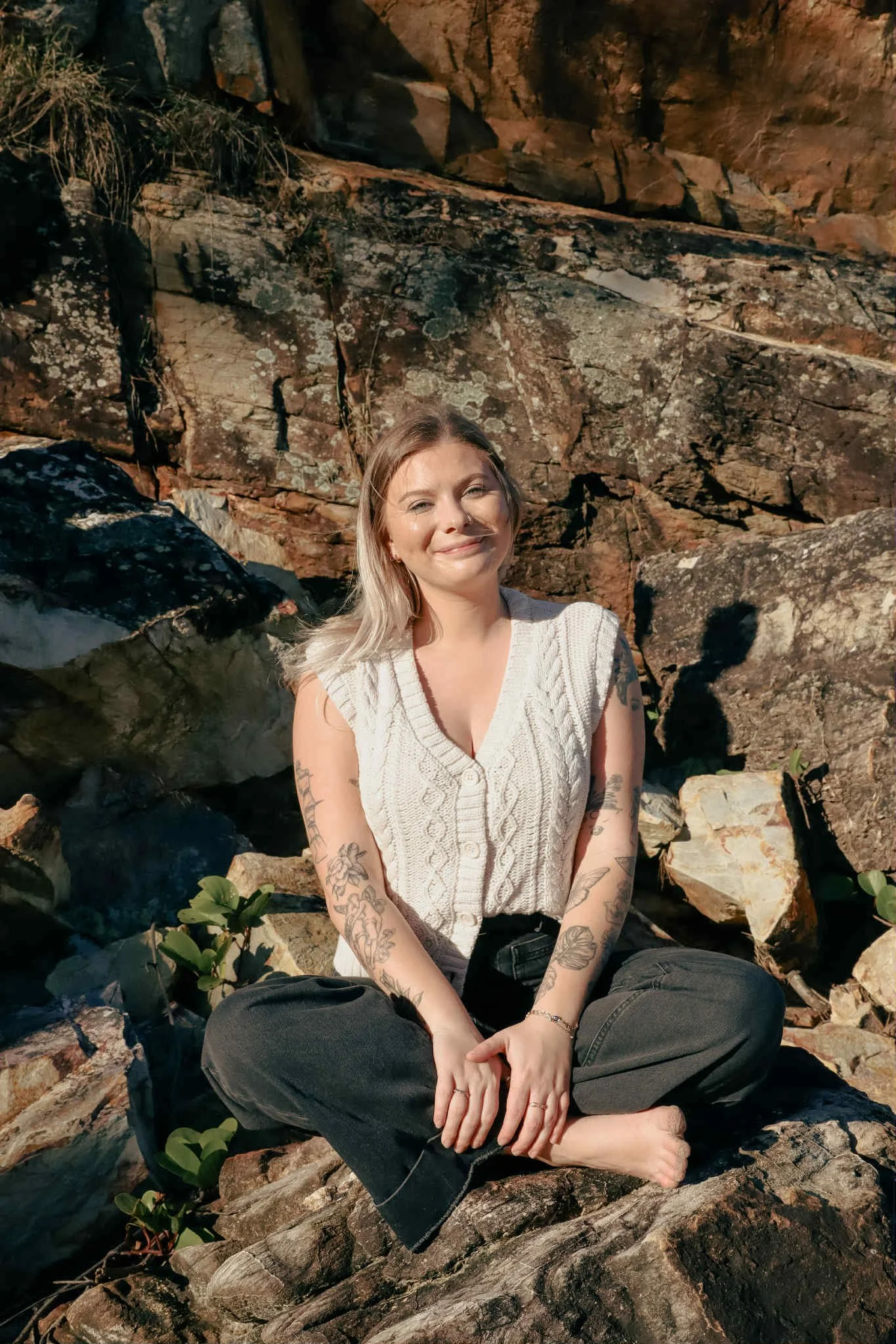 Woman with tattoos sitting cross-legged on rocks near a rocky cliff at sunset.