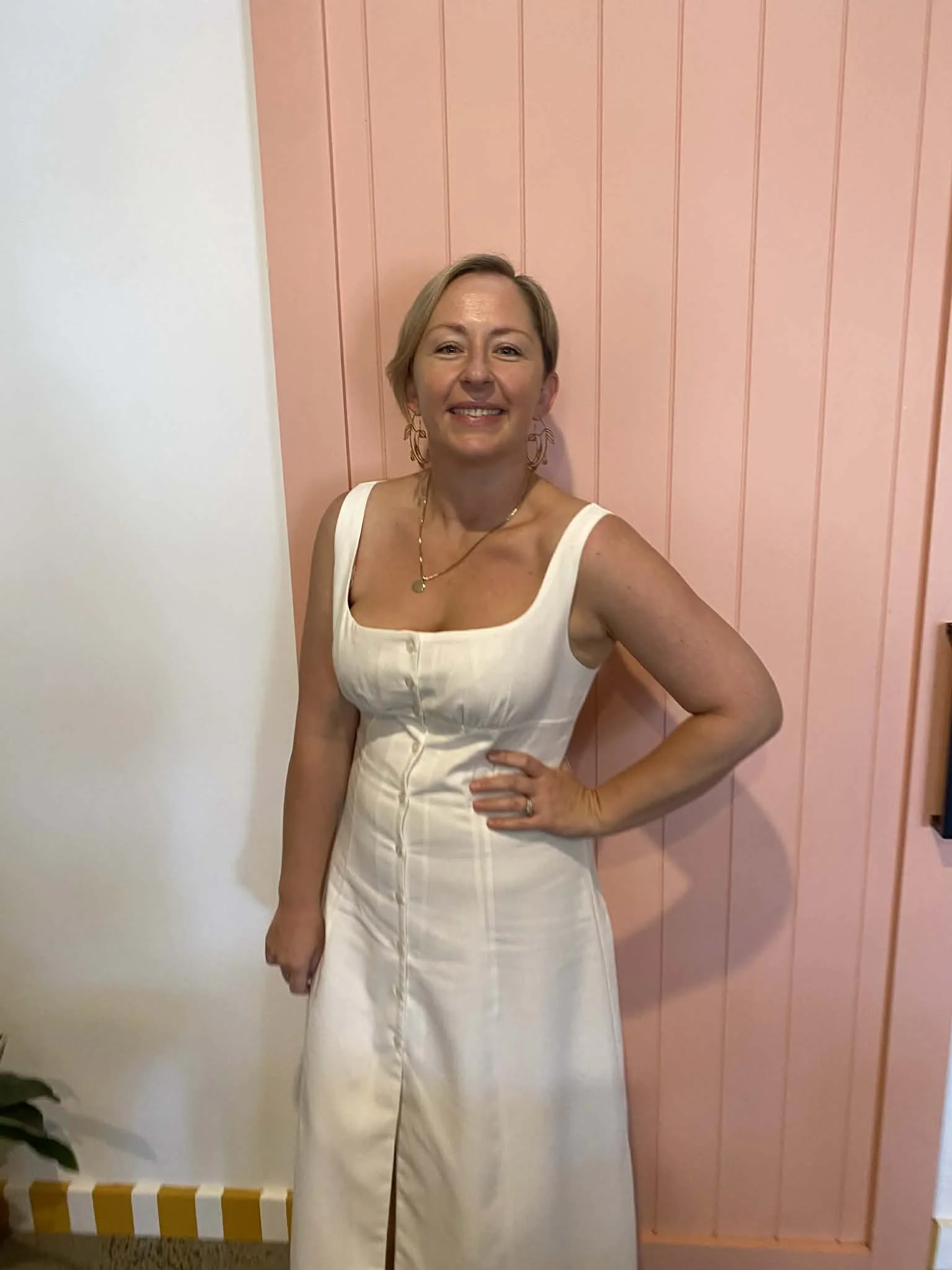 A woman in a white dress standing in front of a pink wooden wall, smiling, with her right hand on her hip.