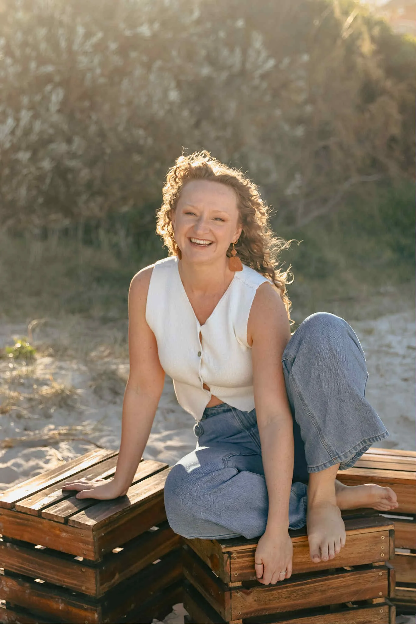 A woman with curly hair smiling, sitting on a wooden bench outdoors with sunlight in the background.