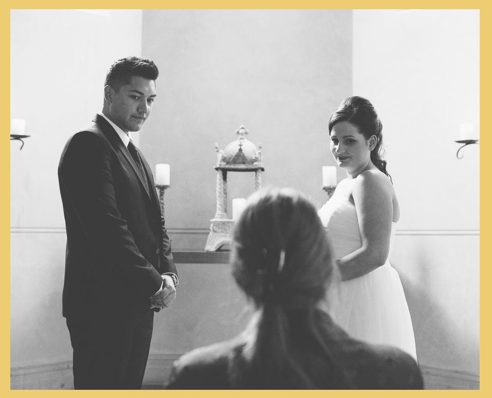 A black-and-white photo of a wedding ceremony with a man in a suit, a woman in a wedding dress, and a person with long hair sitting and facing them.