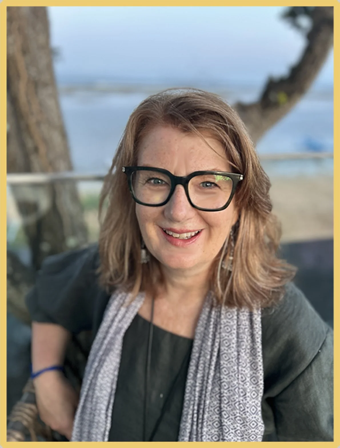 A woman with glasses and earrings smiling outdoors near a body of water and trees.