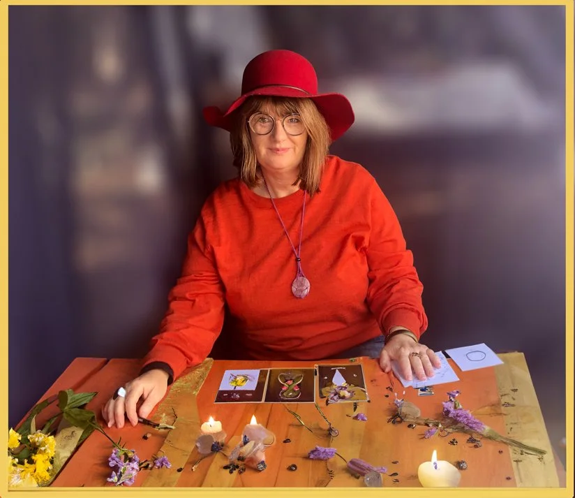 A woman wearing a red hat and orange sweater sitting at a table with tarot cards, candles, flowers, and crystals.
