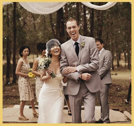 A bride and groom celebrating their wedding outdoors with friends in a wooded area, smiling and laughing.