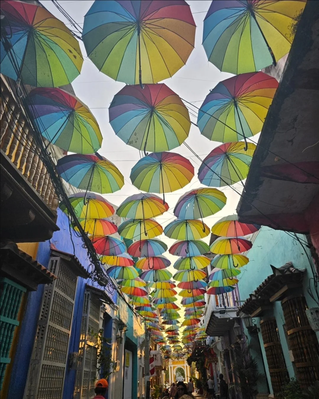 Colorful umbrellas hanging above a narrow street with buildings on both sides and people walking underneath.