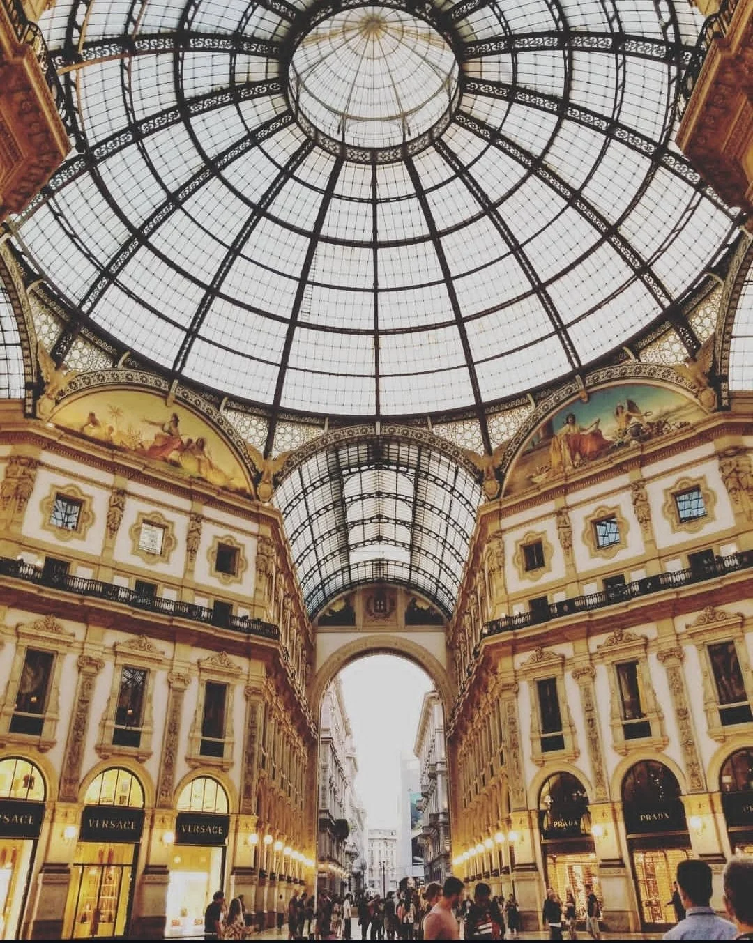 Interior view of a shopping gallery with a glass domed ceiling, ornate architecture, and luxury stores like Versace and Prada, with people walking below.