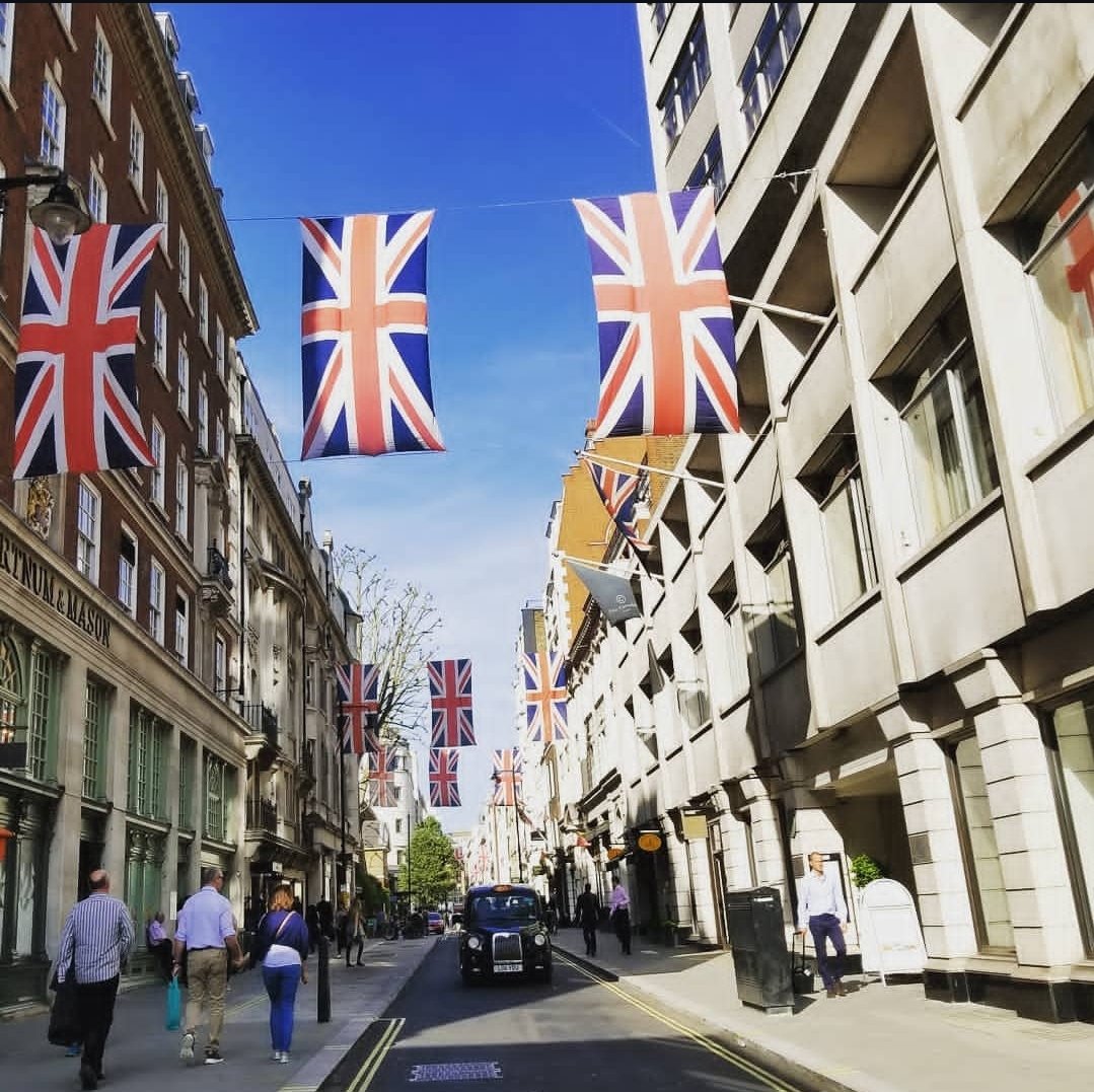 Street scene in London with Union Jack flags hanging across the road, people walking on the sidewalk, and buildings on both sides.