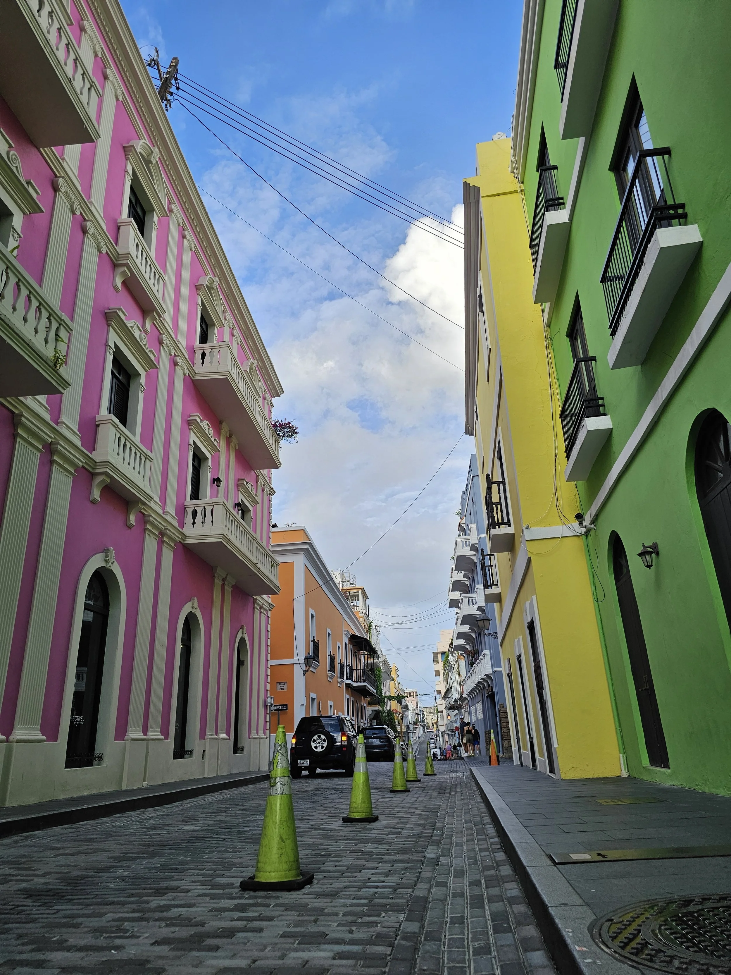 Colorful buildings in a narrow street with parked cars, green traffic cones, and a blue sky with clouds.