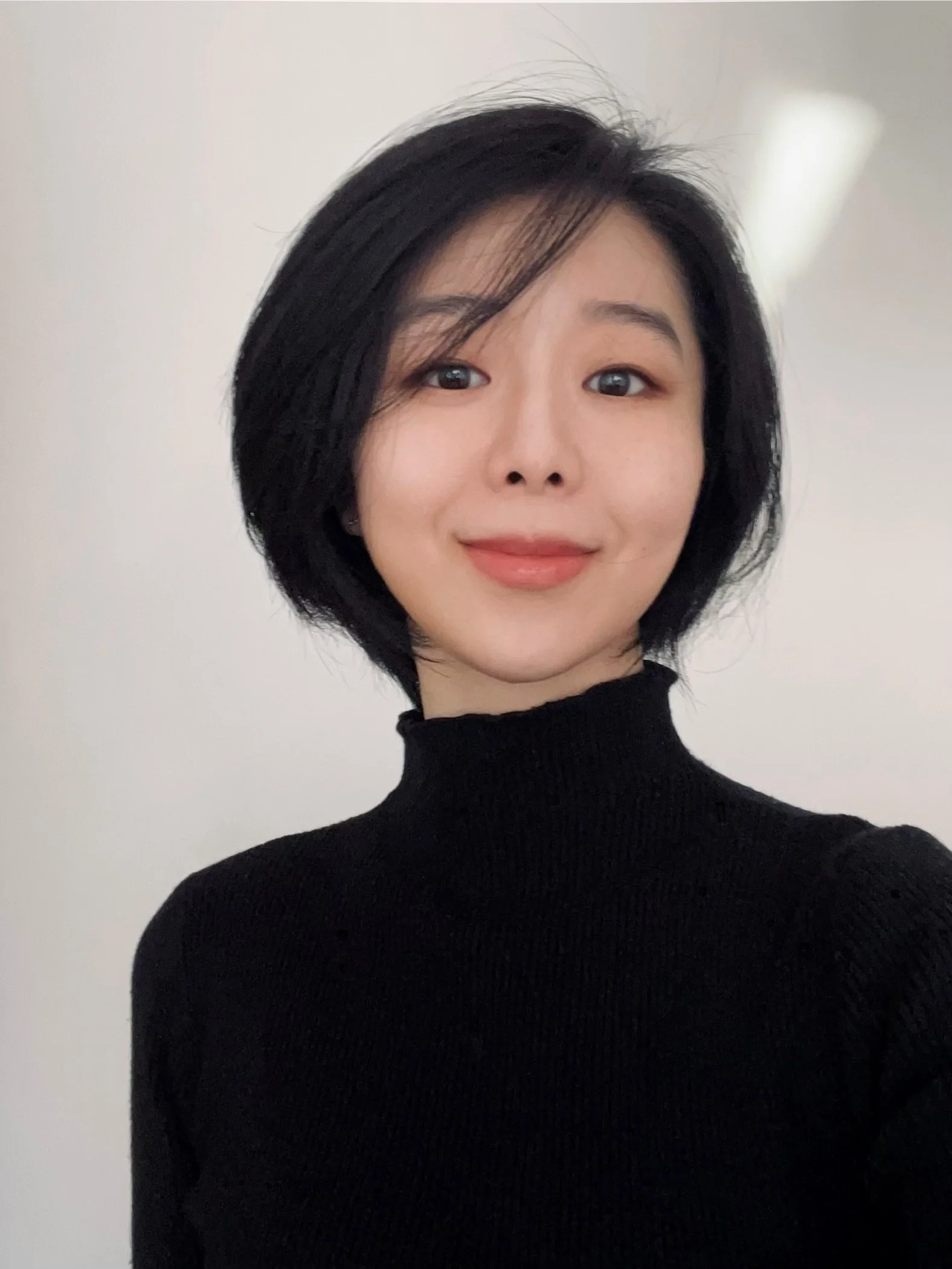 Portrait of a woman with short black hair wearing a black turtleneck, standing against a plain white background.