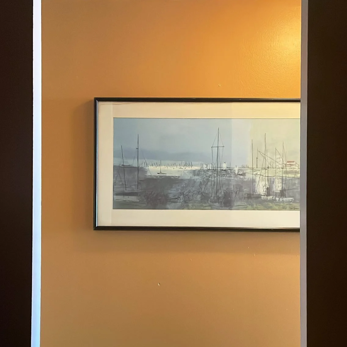 A framed watercolor painting of a harbor scene hangs on a warm mustard-colored wall. The painting depicts several sailboats with tall masts on calm water, with a faint shoreline and distant boats in the background. The frame is thin and black with a 