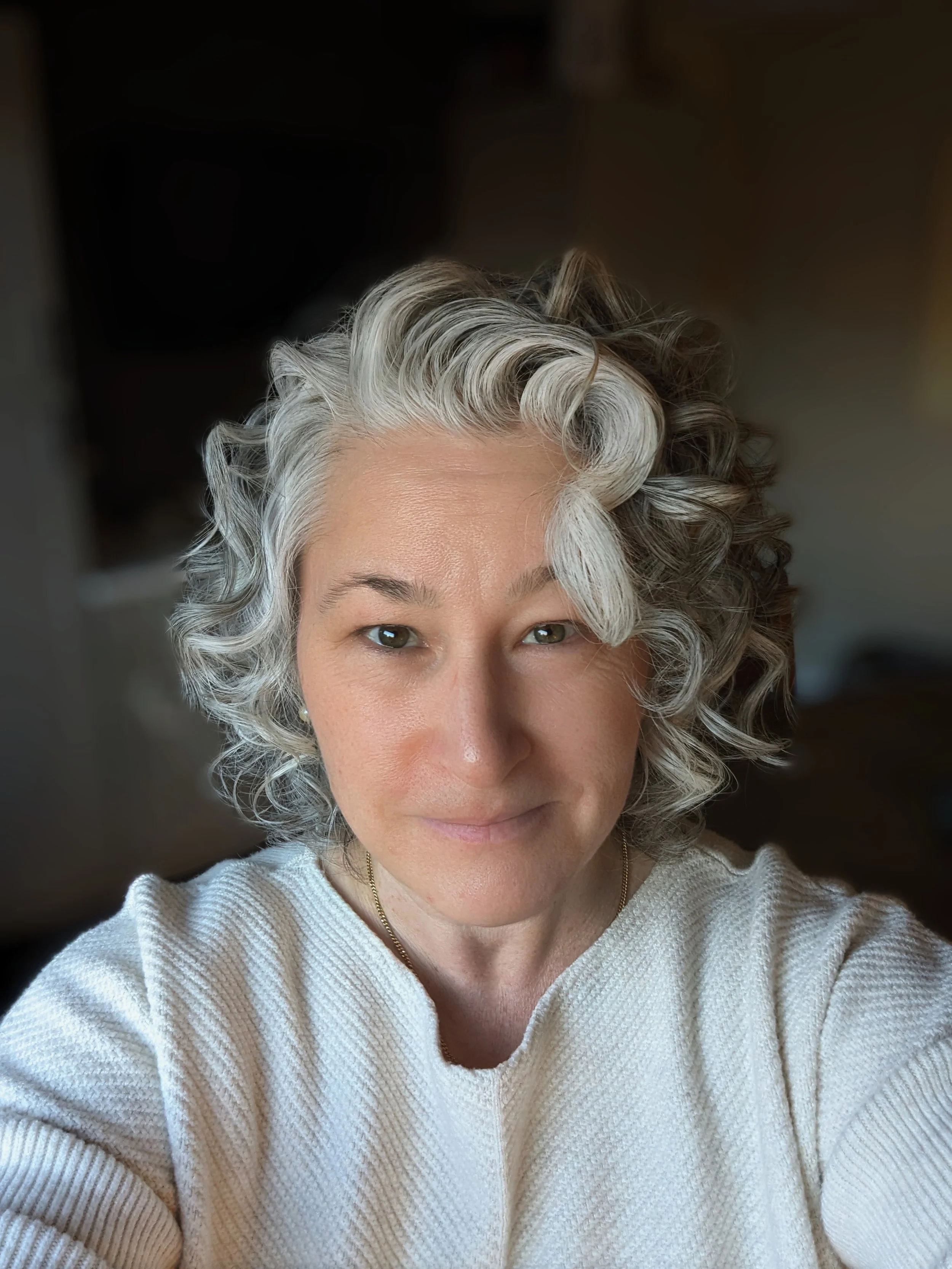 Close-up of a woman with short, curly, silver hair smiling at the camera, wearing a white textured sweater.