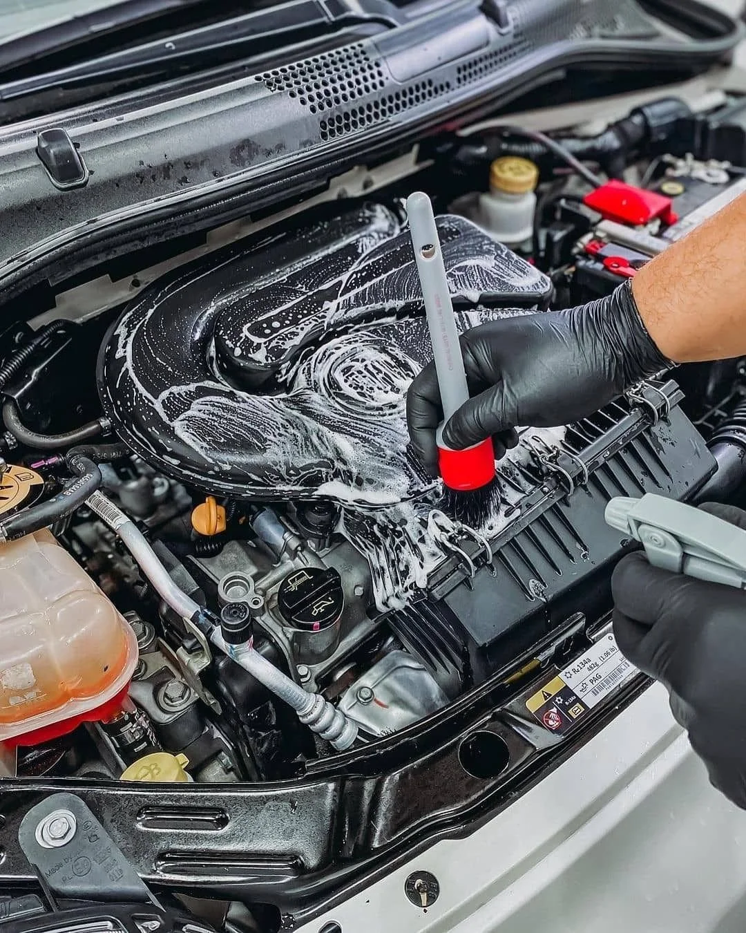 A person wearing black gloves is washing or cleaning a car engine with soap and a brush.