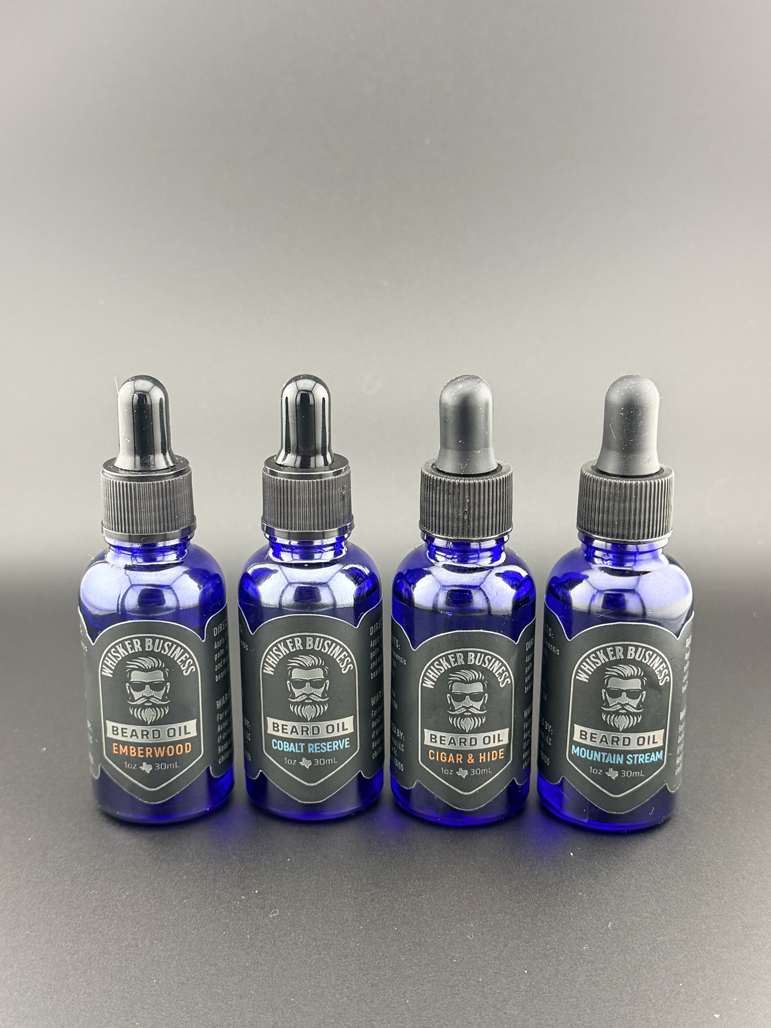Four blue bottles of beard oil with black labels and dropper caps, labeled 'Whisker Business' with different scents: Emberwood, Cobalt Reserve, Cigar & Hide, and Mountain Stream, arranged in a row on a black surface against a gray background.