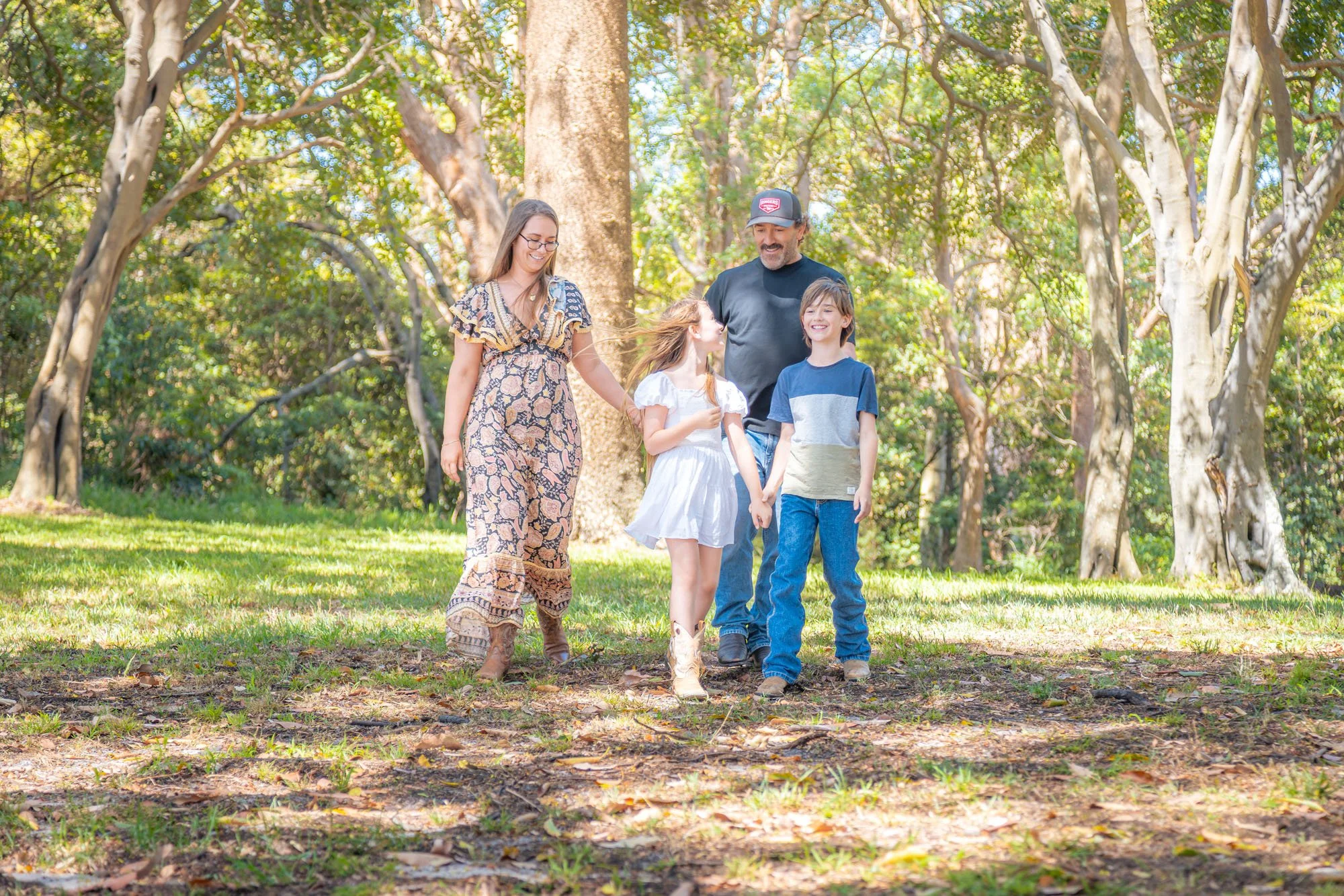A family of four walking in a wooded park, holding hands and smiling on a sunny day during a relaxed outdoor family photoshoot by Southern Exposure Photography.