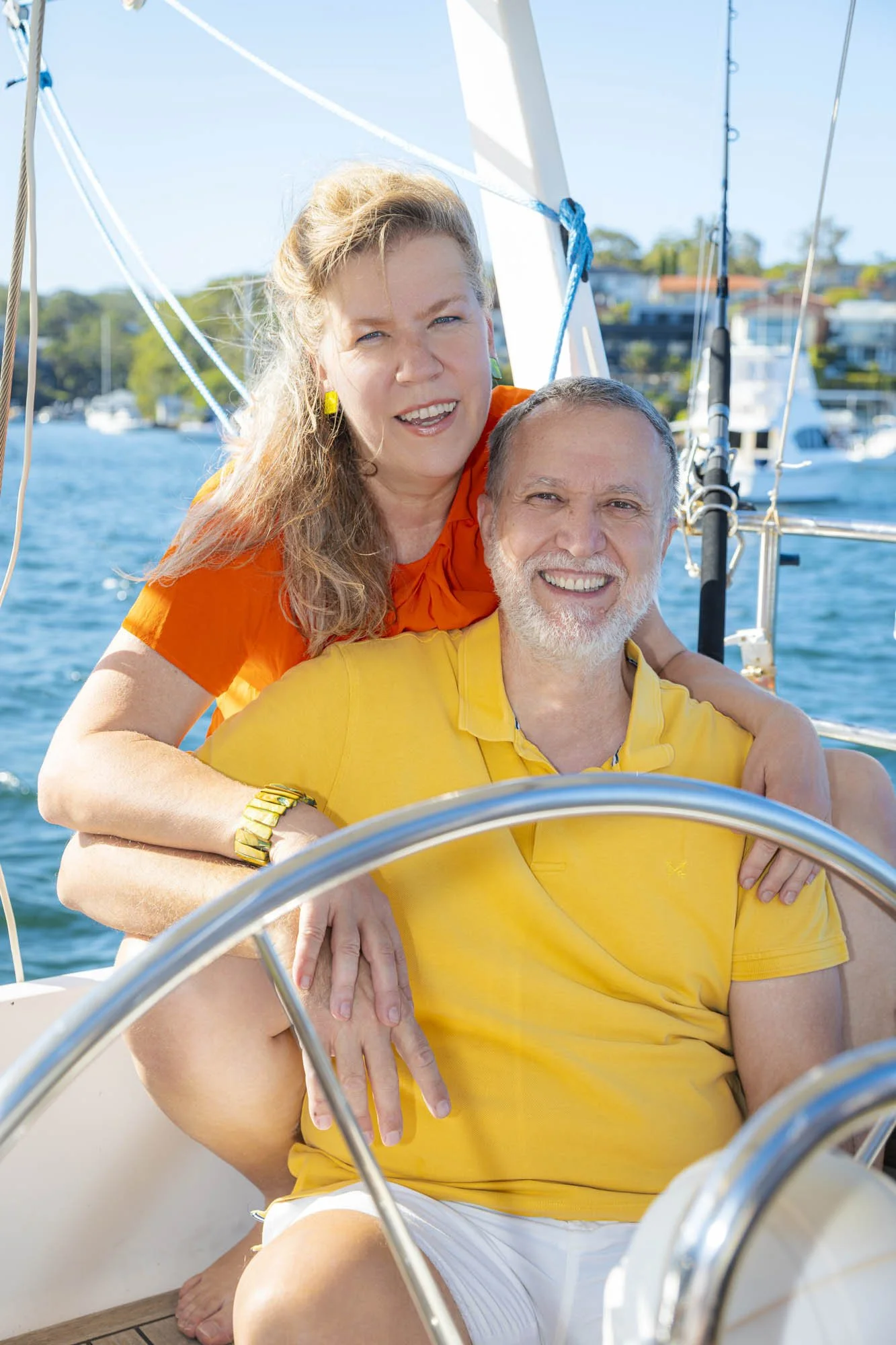 A smiling middle-aged couple on a sailboat, with the woman sitting behind the man and both enjoying a sunny day on the water.
