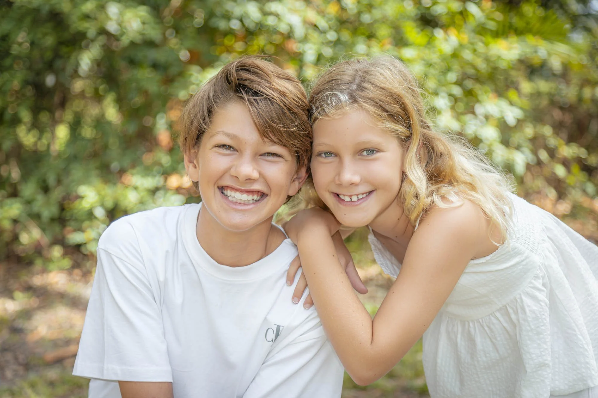 Two children, a boy and a girl, smiling and leaning on each other outdoors surrounded by greenery.