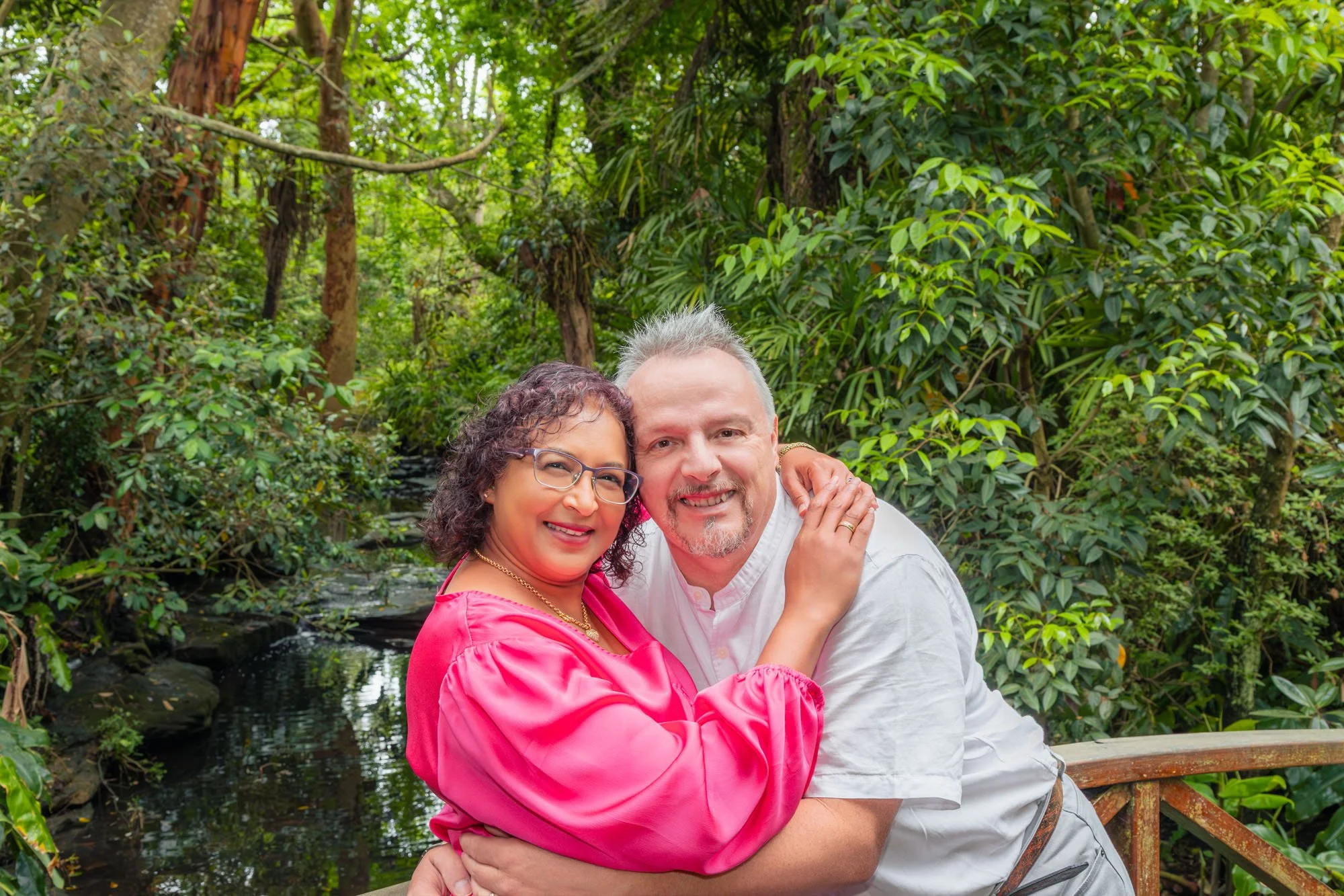 A happy couple hugging each other outdoors near a small pond in a lush green forest during a relaxed outdoor family photoshoot by Southern Exposure Photography.