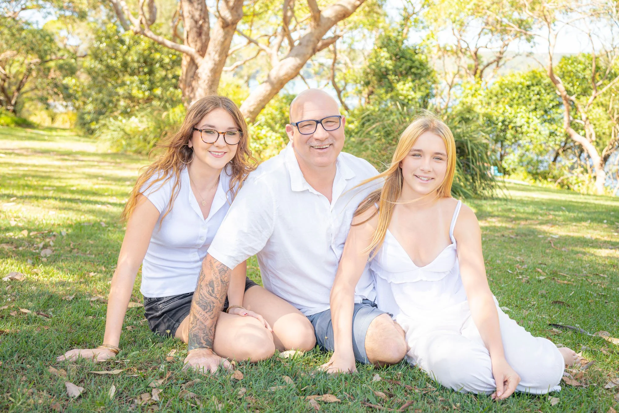 A family of three sitting on grass in a park with trees and blue sky in the background during a relaxed outdoor family photoshoot by Southern Exposure Photography.