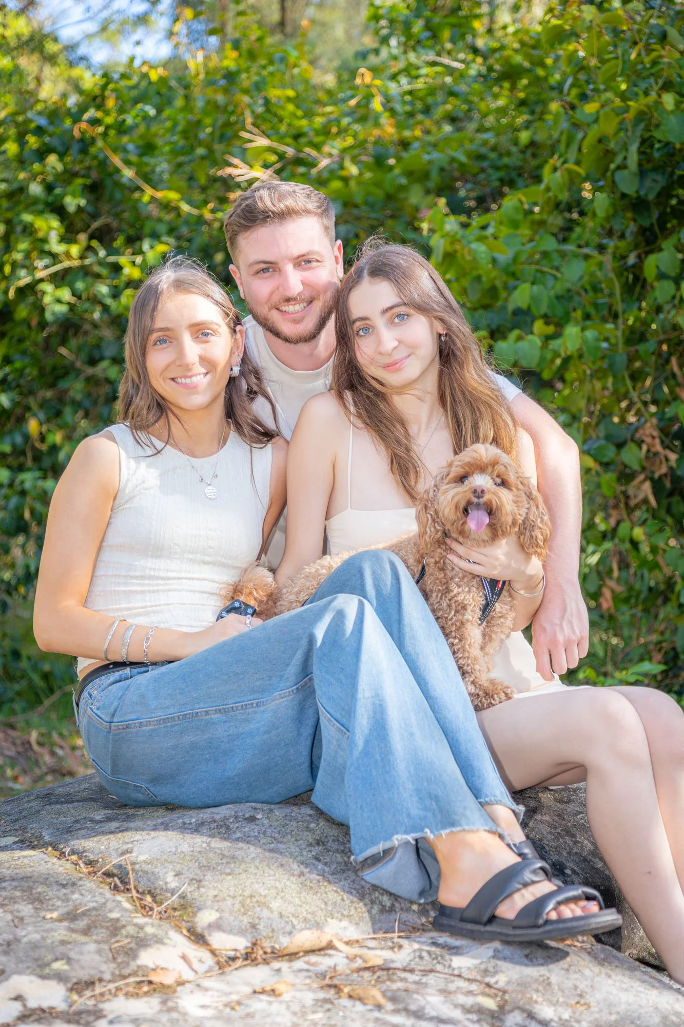A group of siblings, two sisters and one brother, sitting outdoors with a small brown dog, surrounded by lush green foliage, all smiling at the camera during a relaxed outdoor family photoshoot by Southern Exposure Photography.