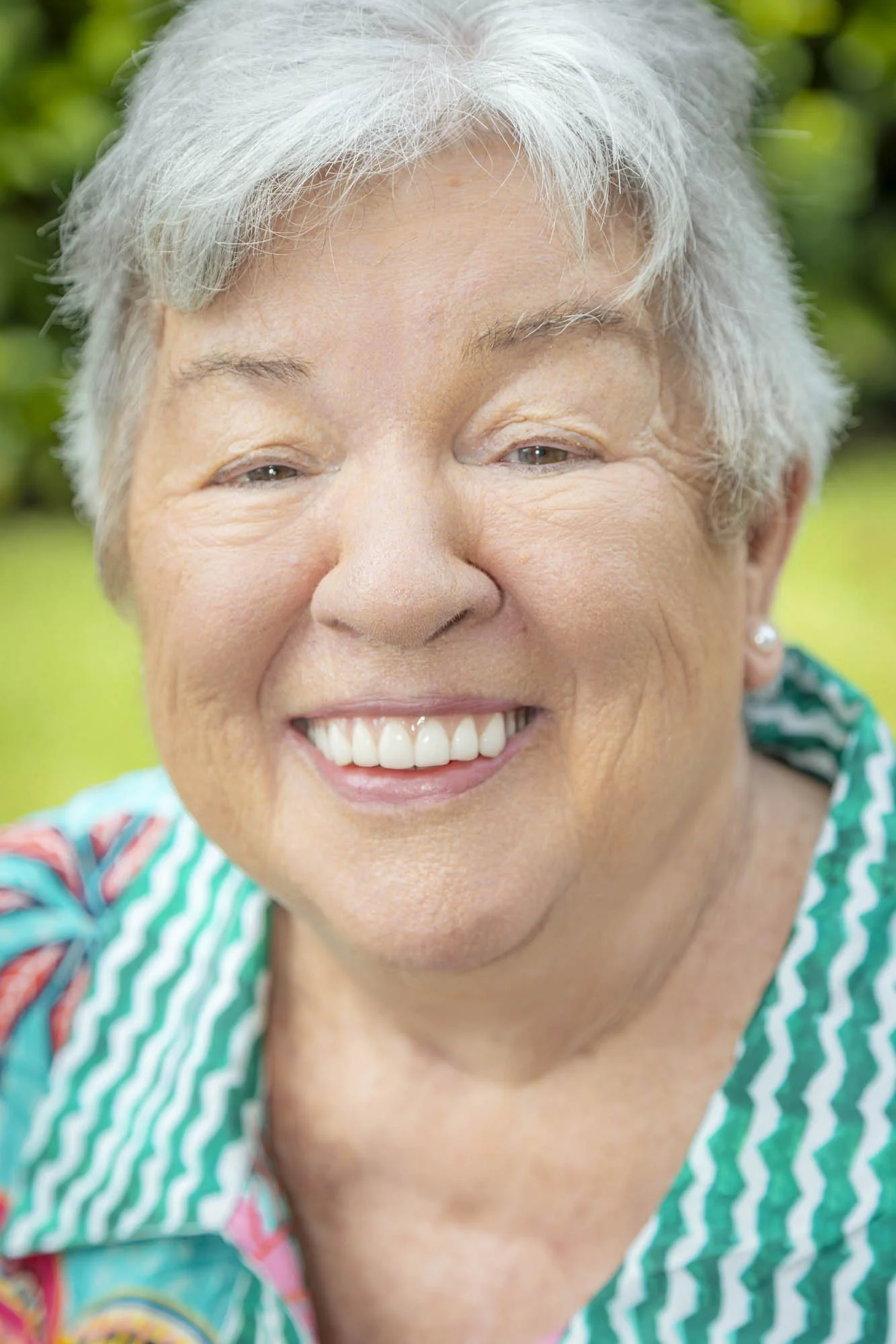 Close-up of an elderly woman with short gray hair smiling in a garden.