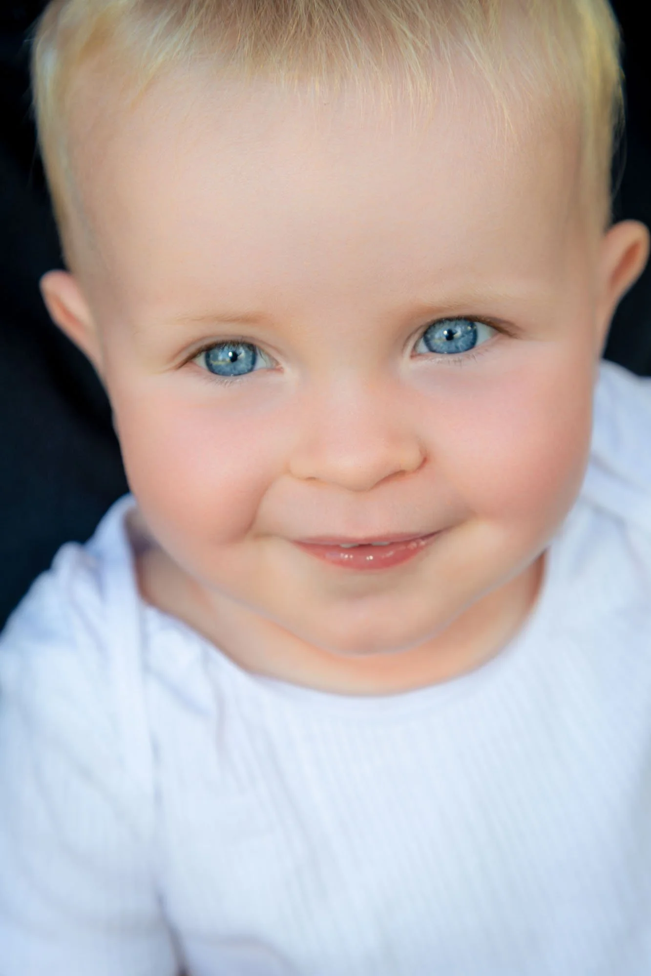 A close-up of a baby boy with bright blue eyes, blonde hair, and a light complexion, smiling while wearing a white shirt during a relaxed outdoor family photoshoot by Southern Exposure Photography.