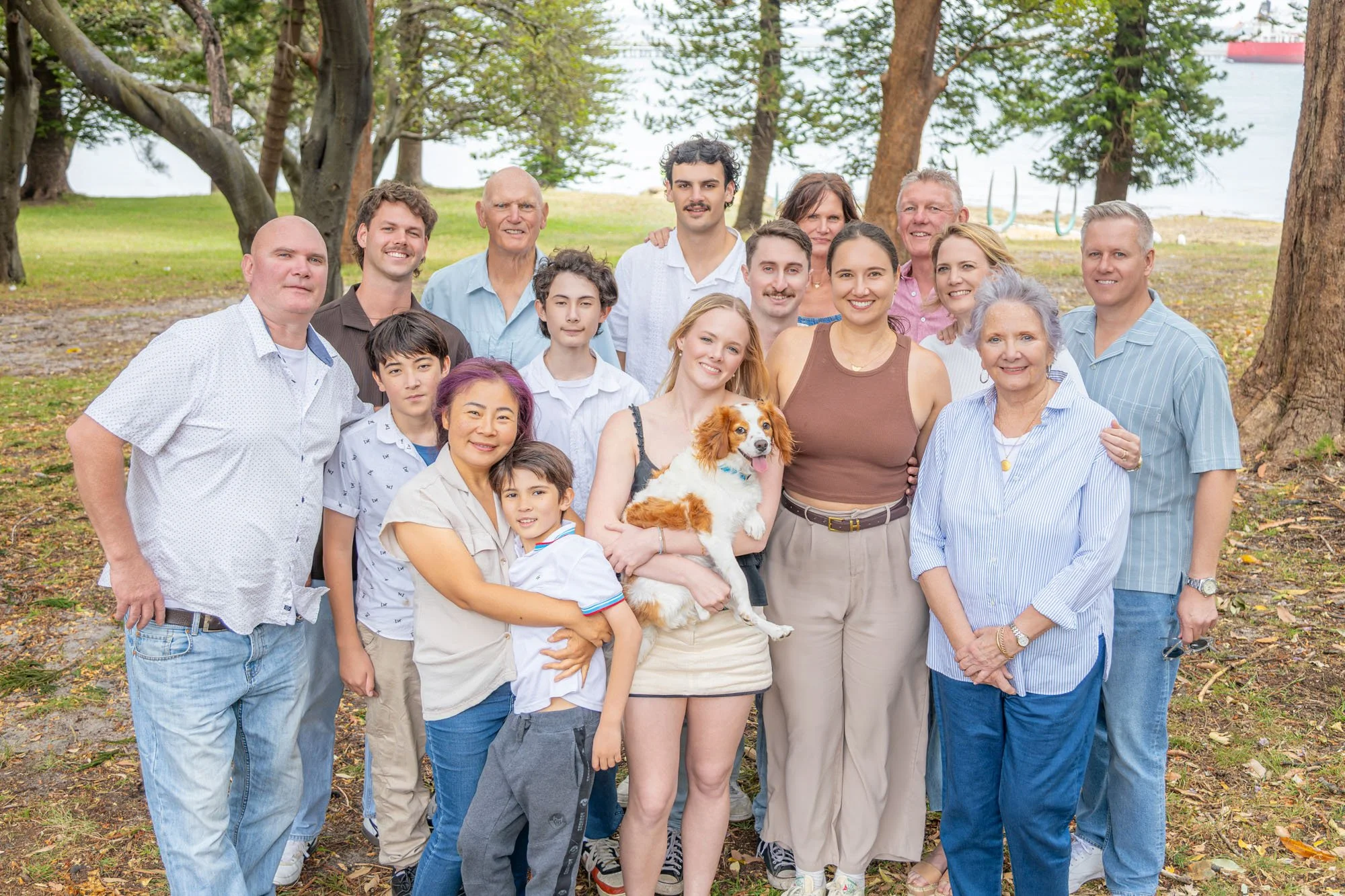 A large multigenerational family group outdoors in a park near water. They are standing together, smiling, with some embracing or holding each other during a relaxed outdoor family photoshoot by Southern Exposure Photography. The group includes men, 