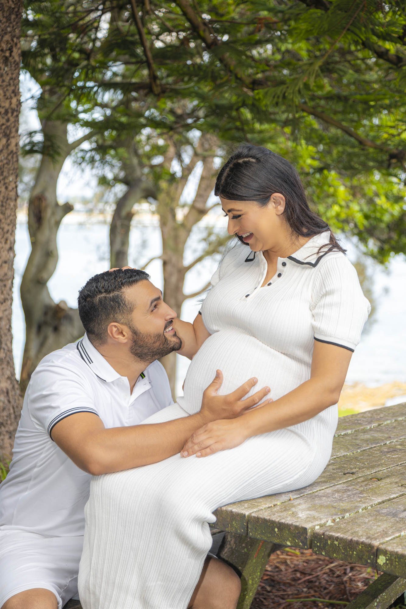 A pregnant woman sitting on a wooden bench outdoors, smiling as she looks down at her partner, who is kneeling and touching her belly, with a background of trees and water.