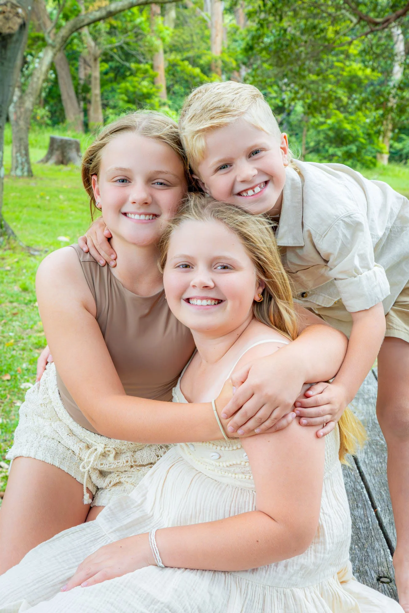 A brother and two sisters smiling and hugging outdoors in a green park with trees during a relaxed outdoor family photoshoot by Southern Exposure Photography.