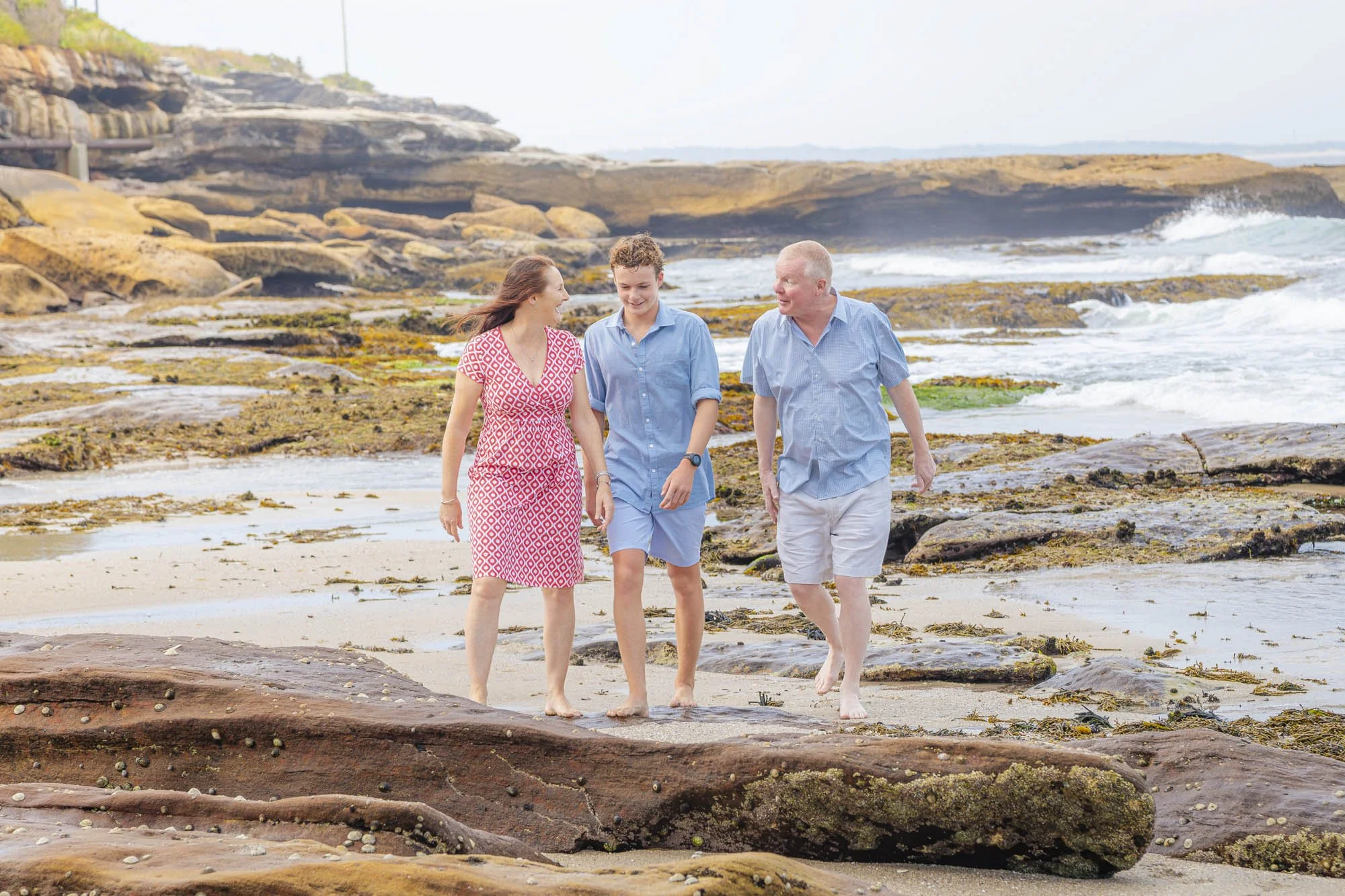 A family of three walking on a rocky beach near the ocean, smiling and enjoying the weather.