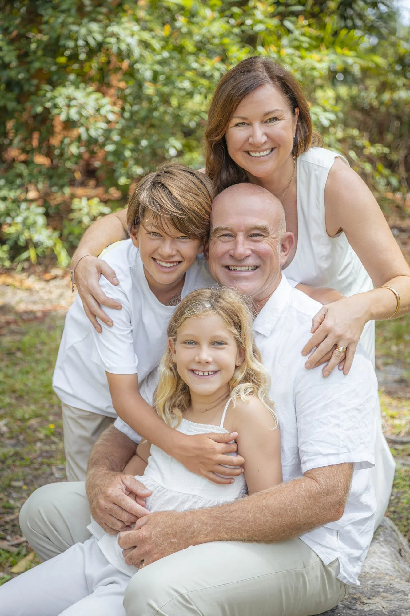 Family of five smiling outdoors, sitting on a log with green foliage in background.