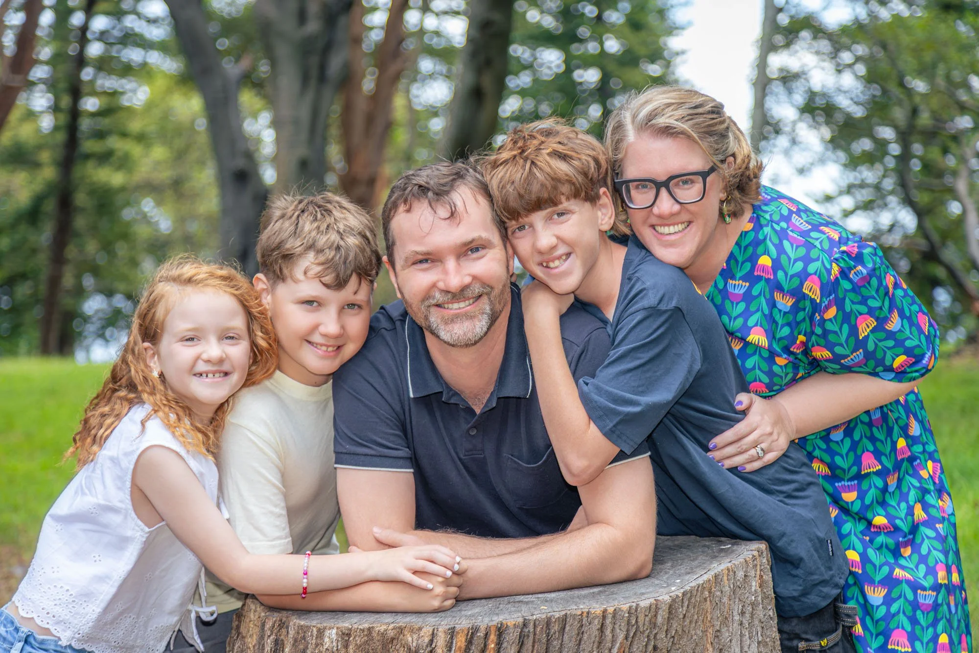 A smiling family of five outdoors, leaning on a tree stump with trees and green grass in the background during a relaxed outdoor family photoshoot by Southern Exposure Photography.