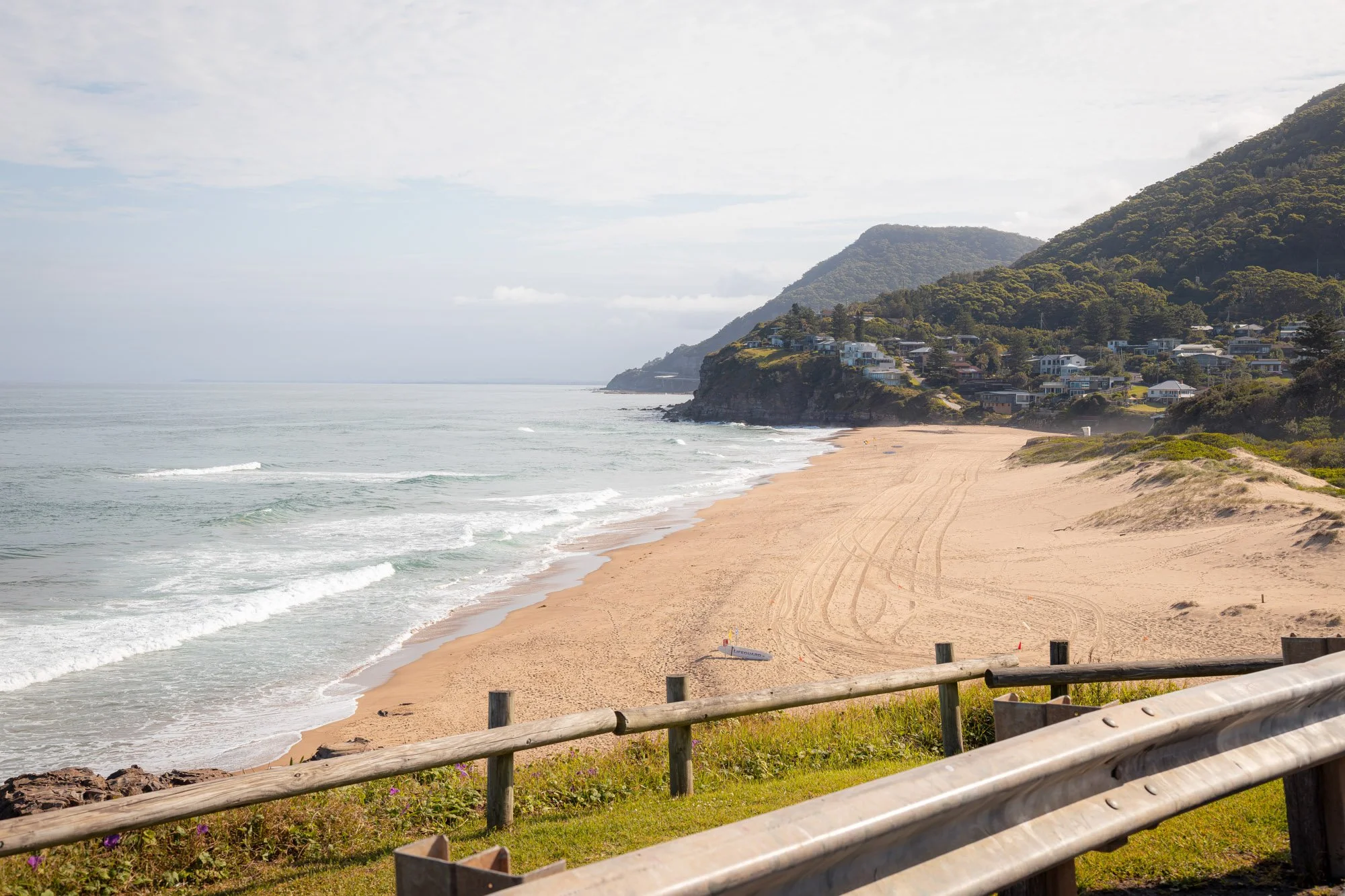 An image of Stanwell Park Beach. It features a view of a sandy beach with gentle waves, a grassy area with a wooden fence, houses on a hillside, and mountains in the background under a partly cloudy sky.