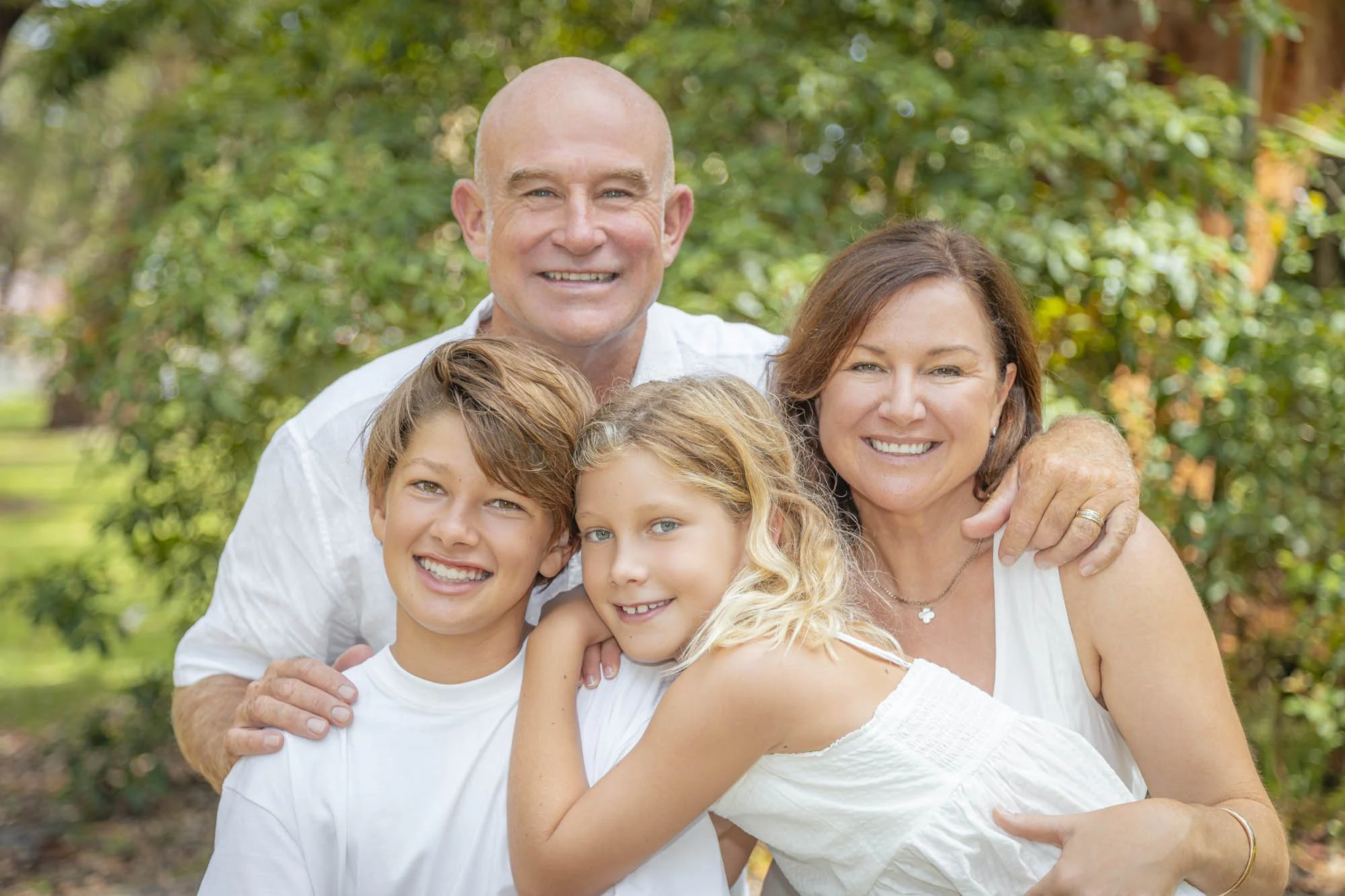 A smiling family of four in a park, with a bald man, a woman with brown hair, a boy, and a girl, all wearing white clothing, close together and embracing.