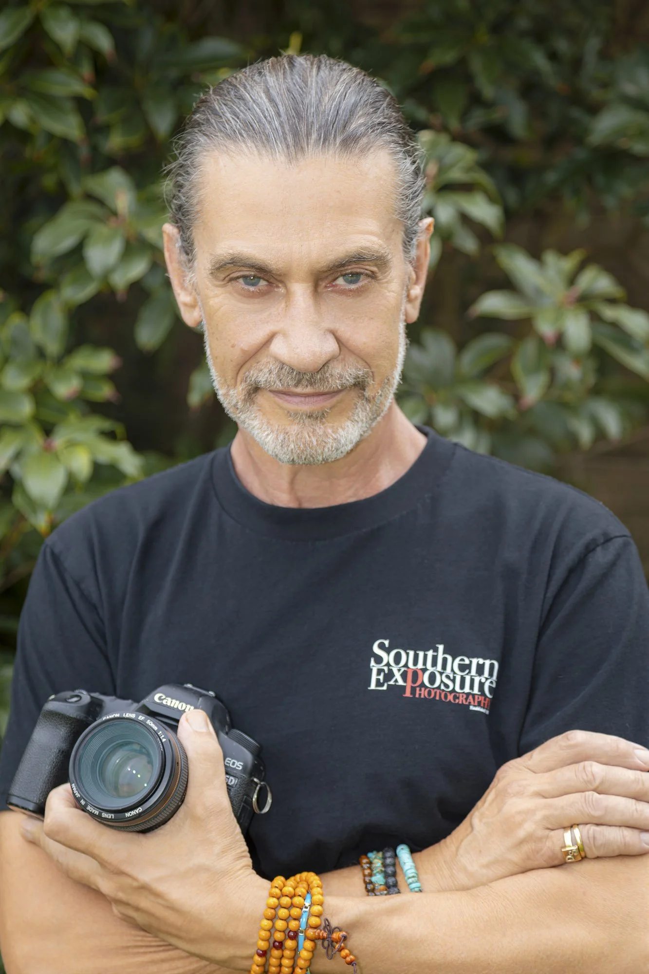 A middle-aged man with slightly grey hair and a beard standing outdoors in front of greenery, holding a Canon DSLR camera, wearing a black T-shirt with 'Southern Exposure Photography' logo, and colorful beaded bracelets on his wrist.