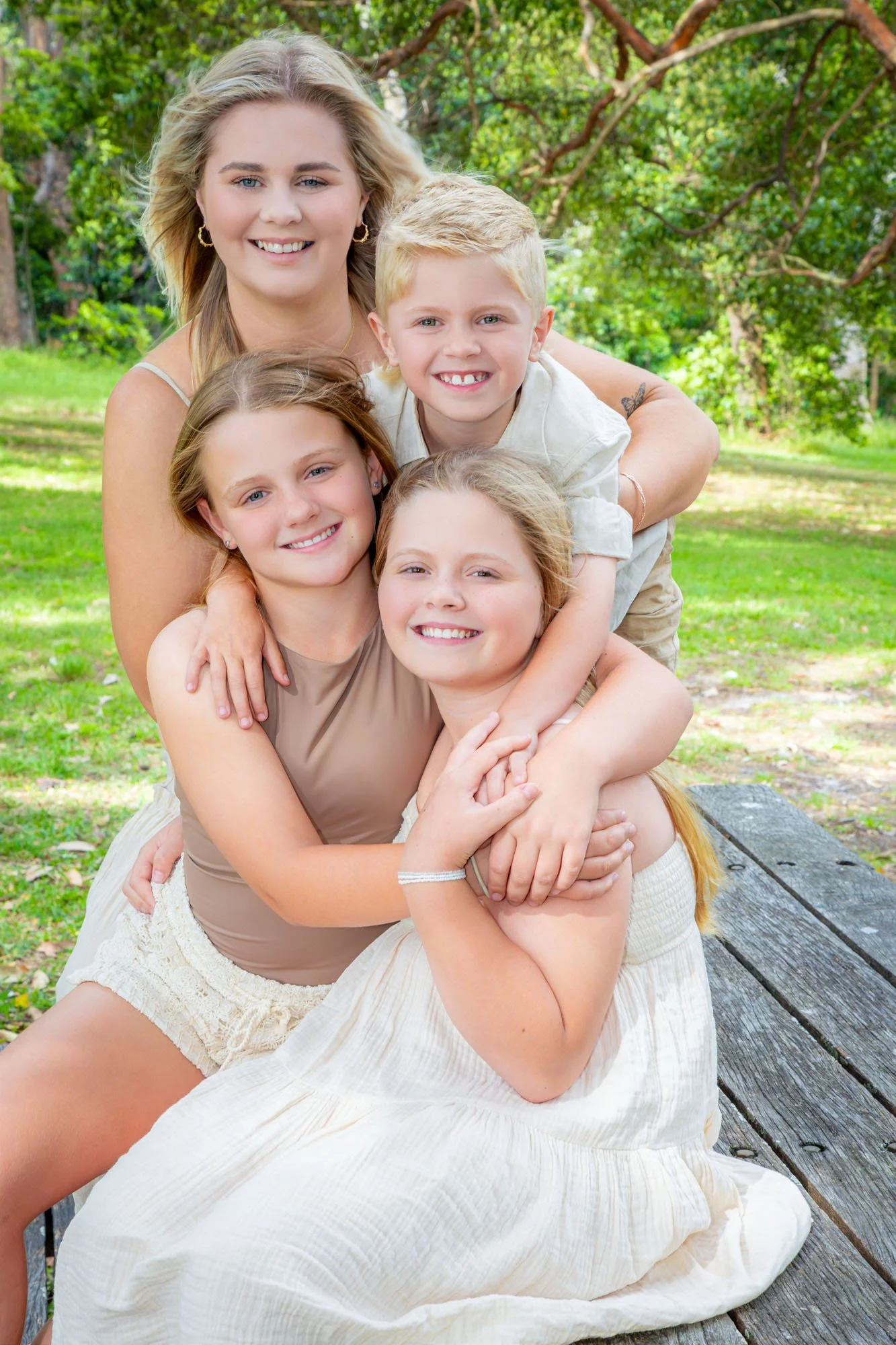 A smiling mother and four children, three girls and one boy, sitting on a wooden bench outdoors in a park with green trees and grass in the background during a relaxed outdoor family photoshoot by Southern Exposure Photography.
