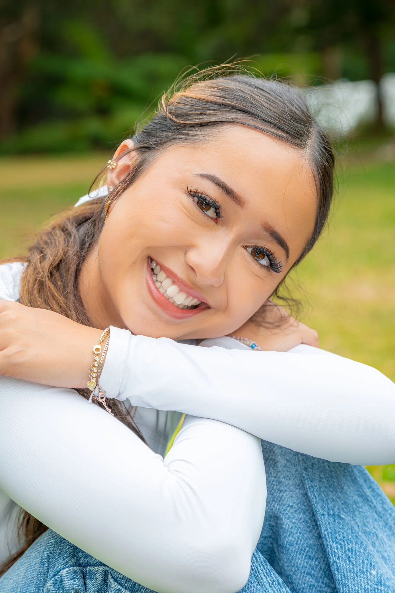 A young woman smiling happily, resting her head on her crossed arms outdoors. She has light brown hair, brown eyes, and is wearing a white long-sleeve top with jewellery during a relaxed outdoor family photoshoot by Southern Exposure Photography. The