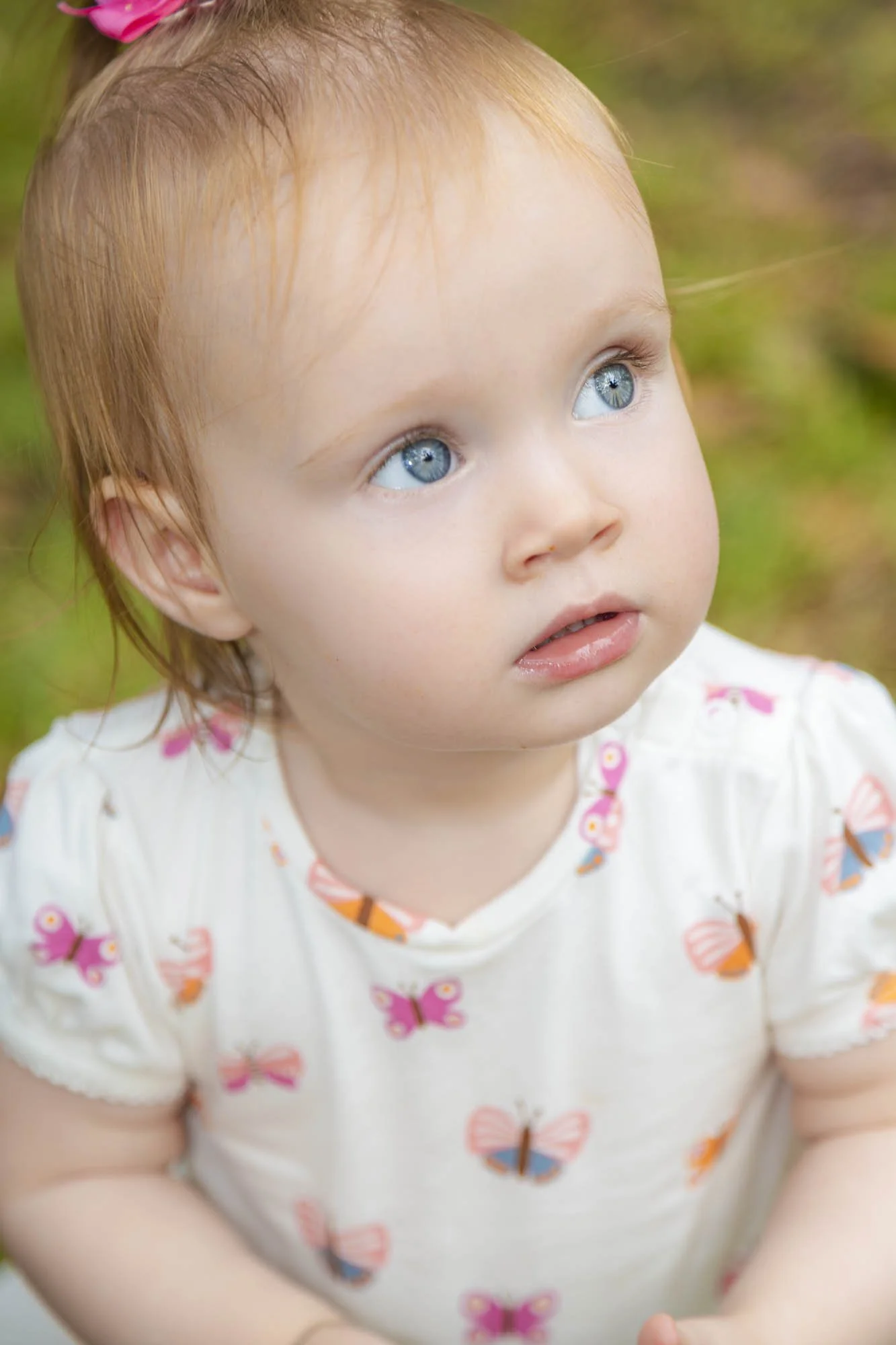 Close-up of a young girl with red hair, blue eyes, and a white shirt with pink, orange, and blue butterfly prints, outdoors on a grassy background.