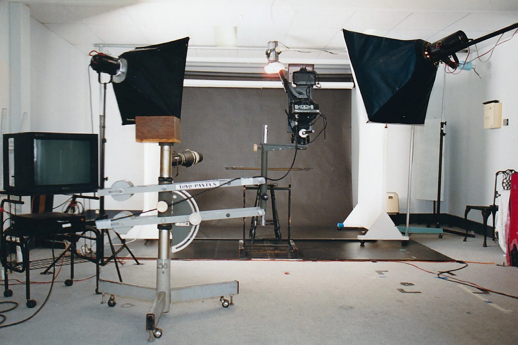A photography studio with professional lighting on stands, a camera setup on a tripod, monitor, and a backdrop in a room with a beige carpet and white walls.