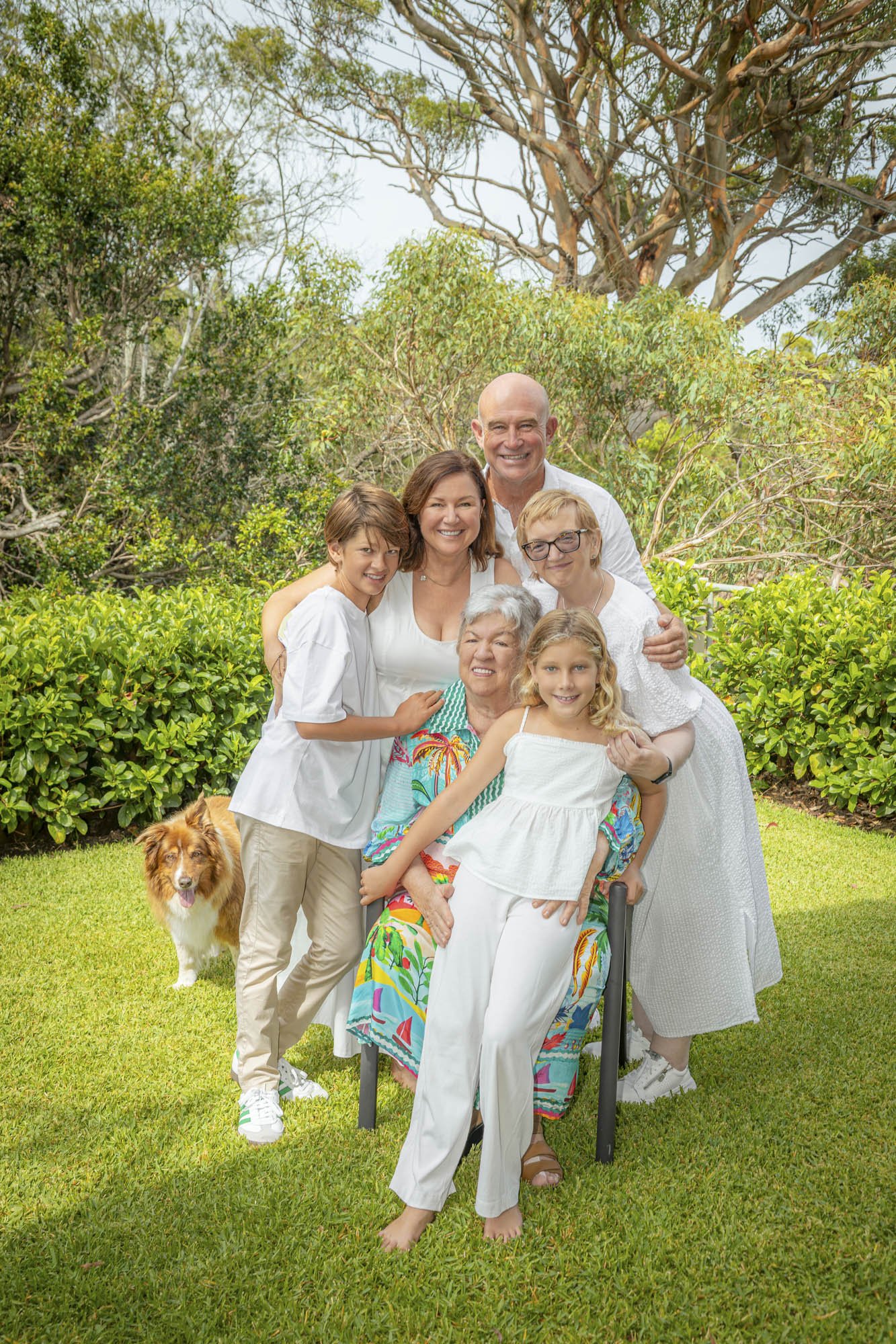A multigenerational family of eight people and a dog gathered outdoors in a lush garden, smiling at the camera.
