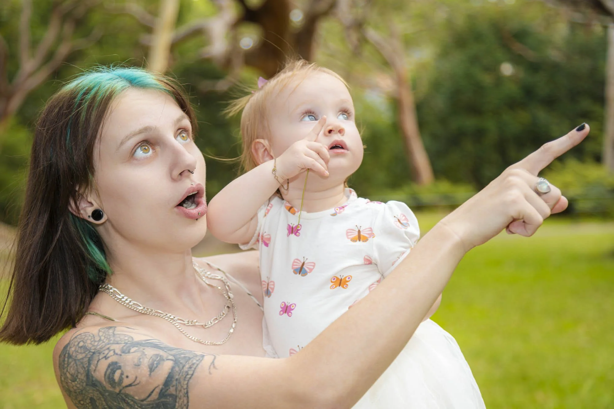 A young woman with dark hair and tattoos on her arm holds a child in a park, both looking up and pointing at something in the sky.